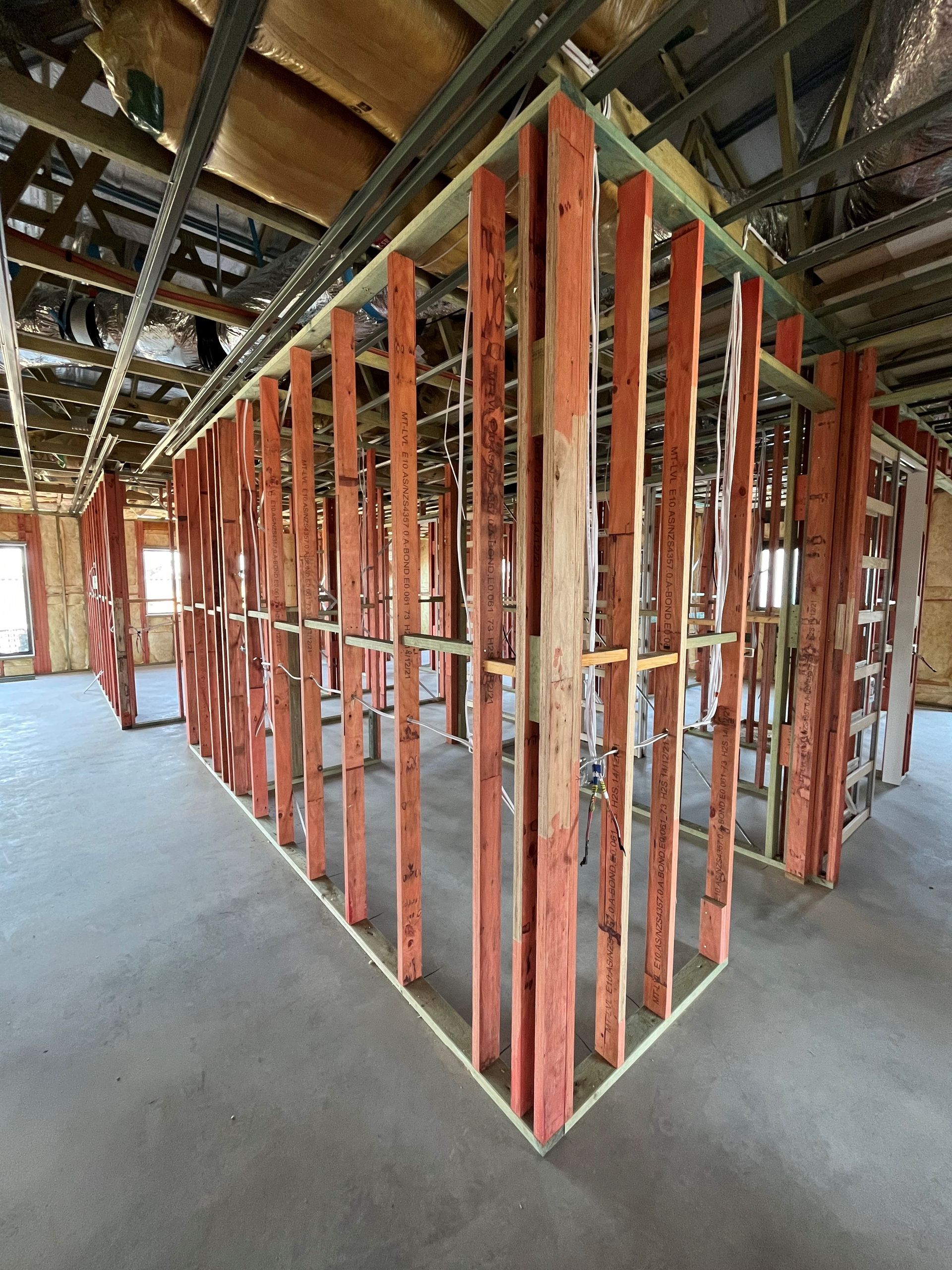 Interior framing of a building under construction with exposed wooden studs, ceiling beams, and a concrete floor.