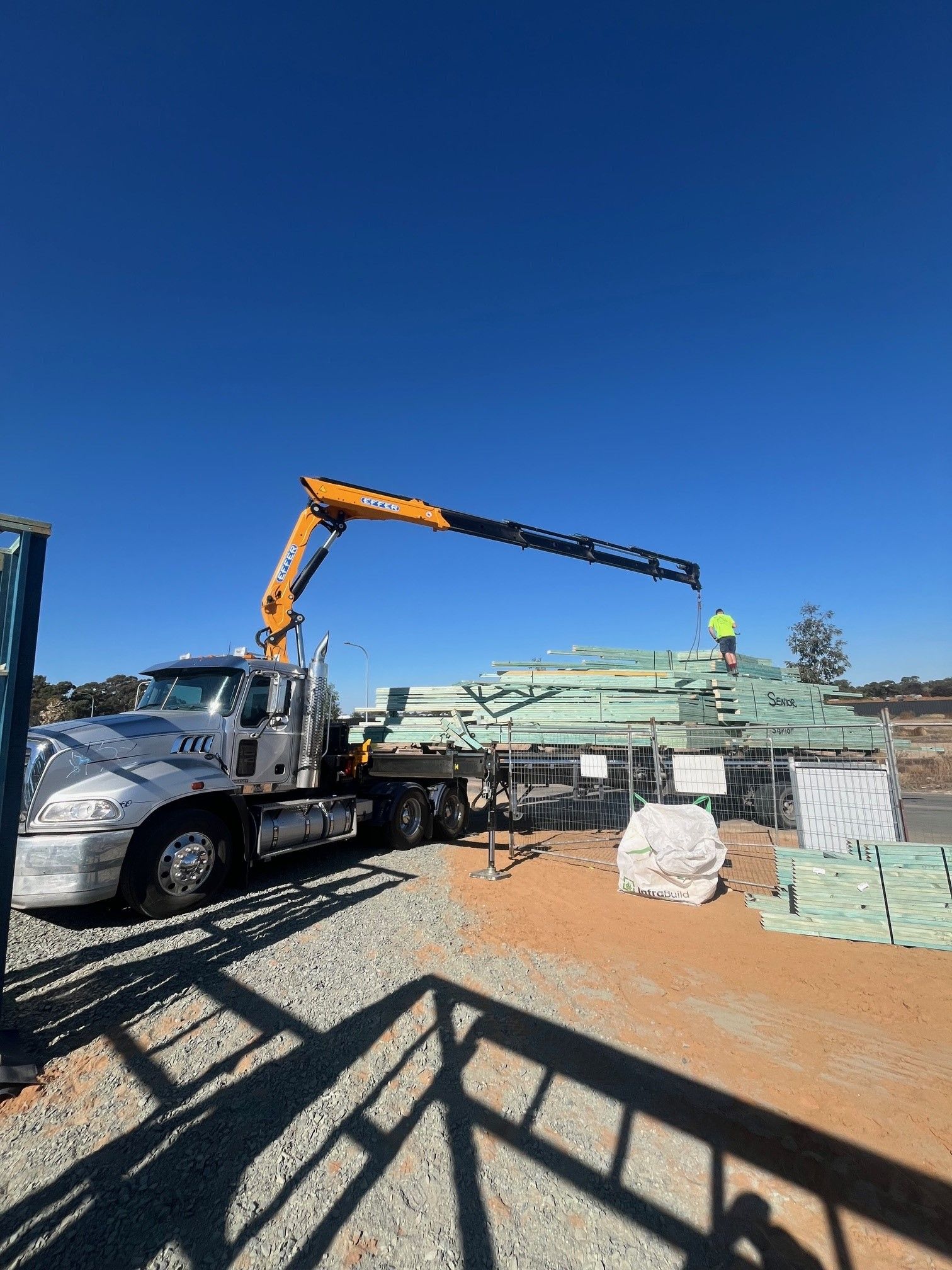 A truck with an extended crane unloading lumber at a construction site under a clear blue sky.