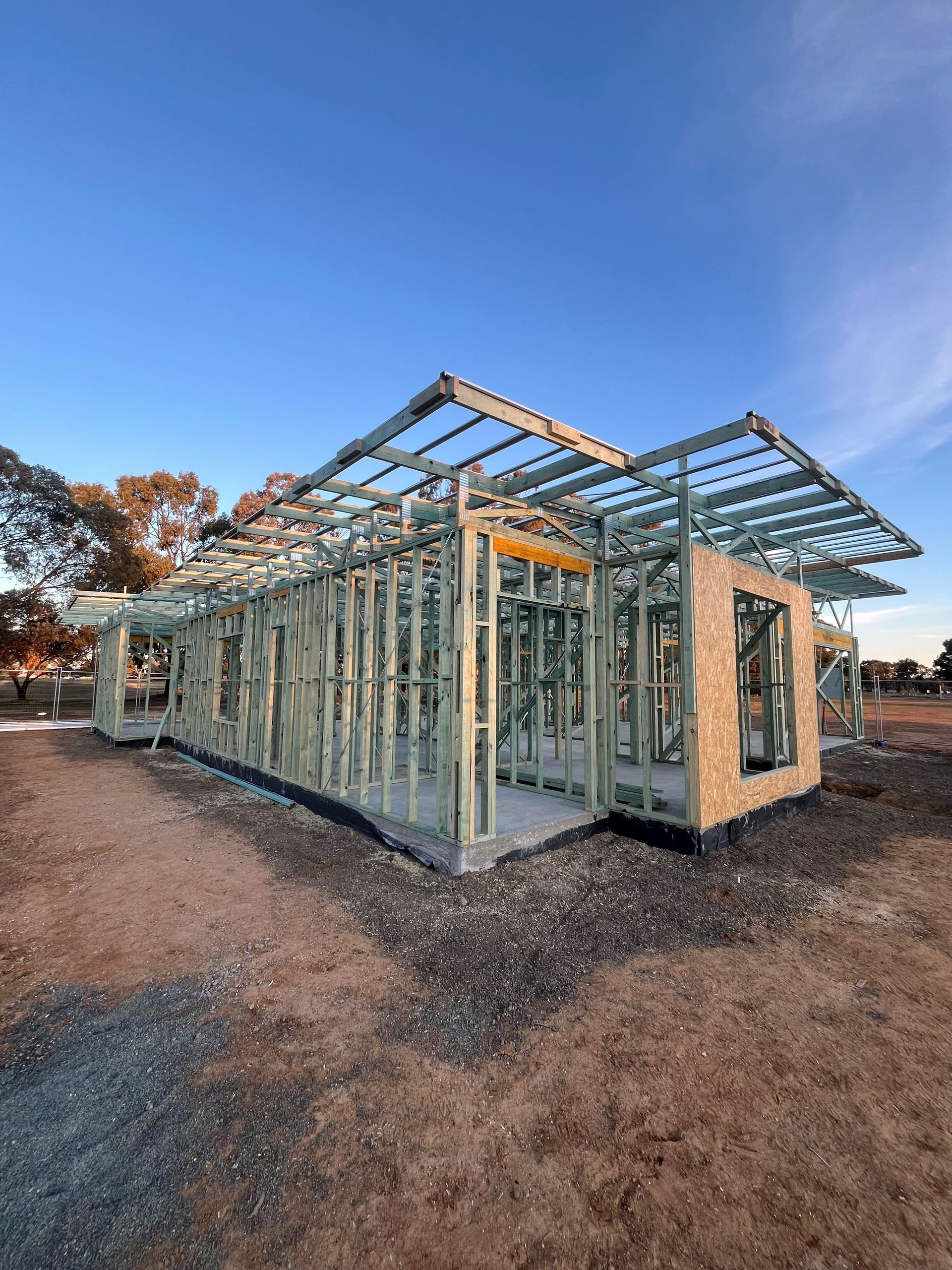 House frame under construction, showcasing wooden studs and roof beams against a blue sky. The setting is a gravel lot.