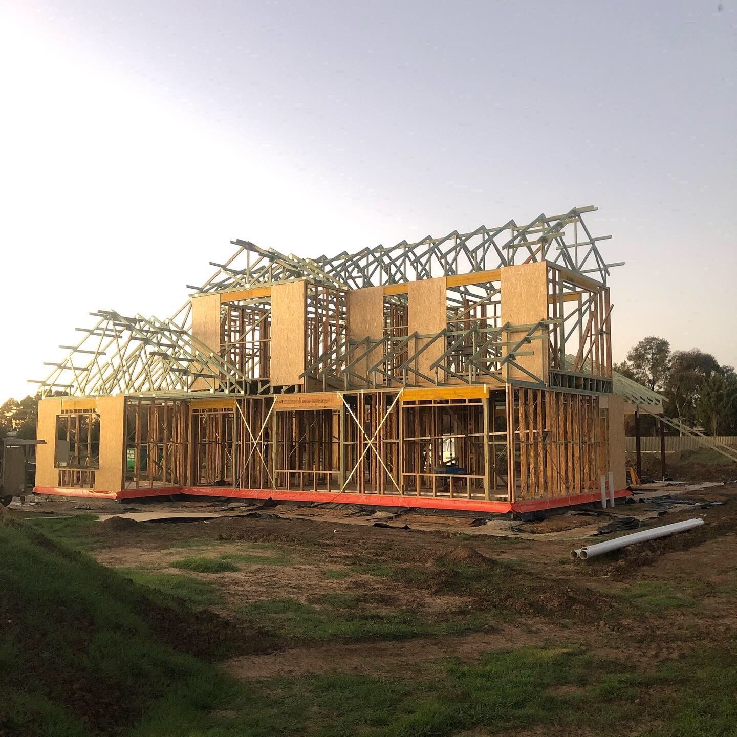 Construction of a two-story house with wooden framing and plywood sheathing, set in a rural landscape under a dusky sky.