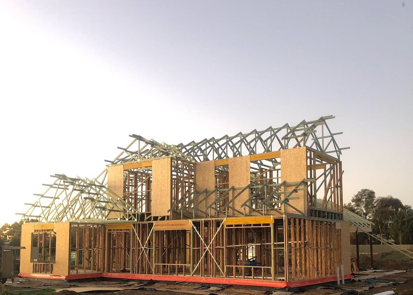 Wooden frame of a two-story house under construction with exposed roof trusses, set against a pale blue sky.