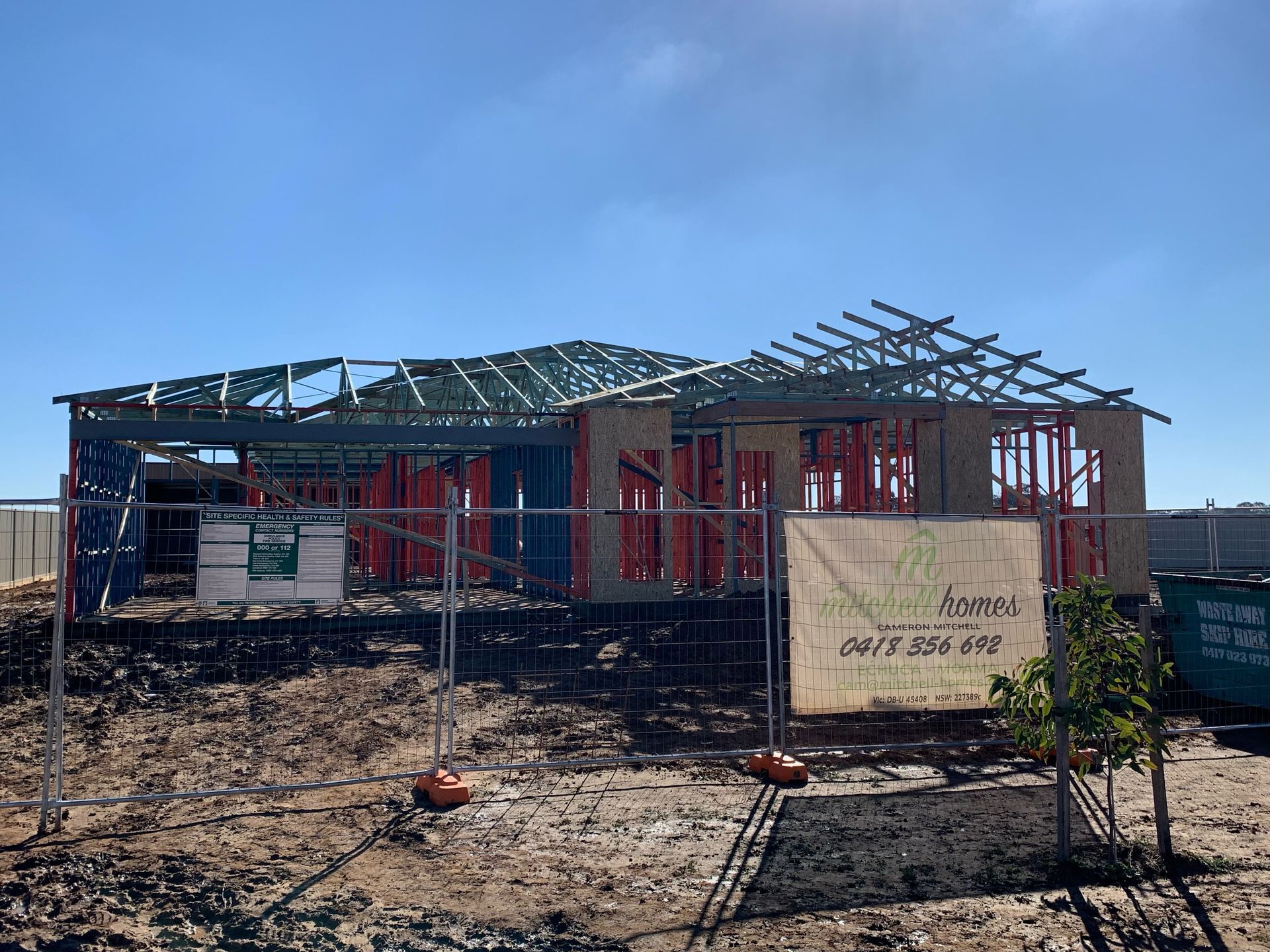 A house under construction; framing and roof beams visible against a clear blue sky. Construction fencing surrounds the site.