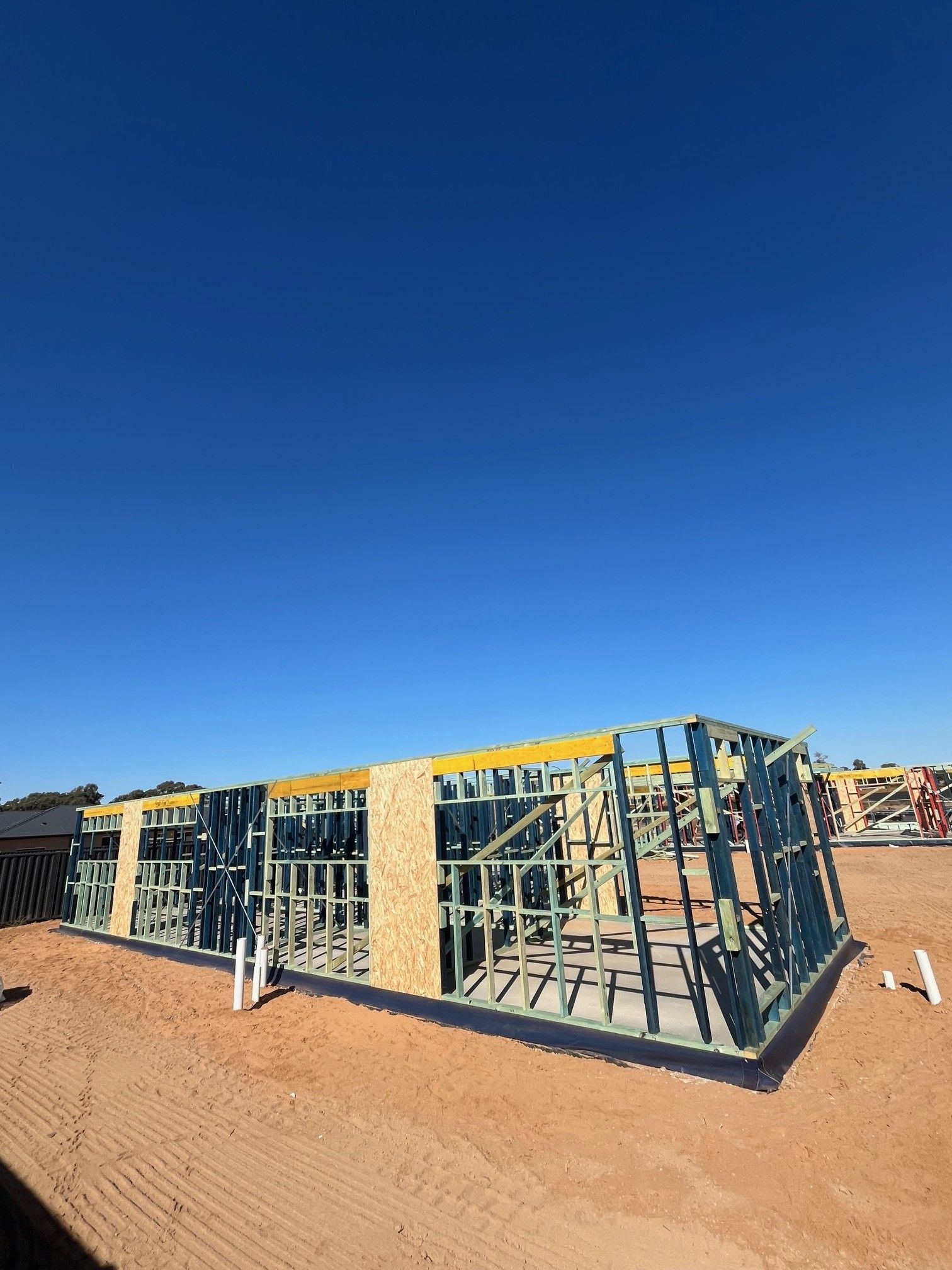 Wooden frame of a building under construction on a sandy lot, against a clear blue sky.