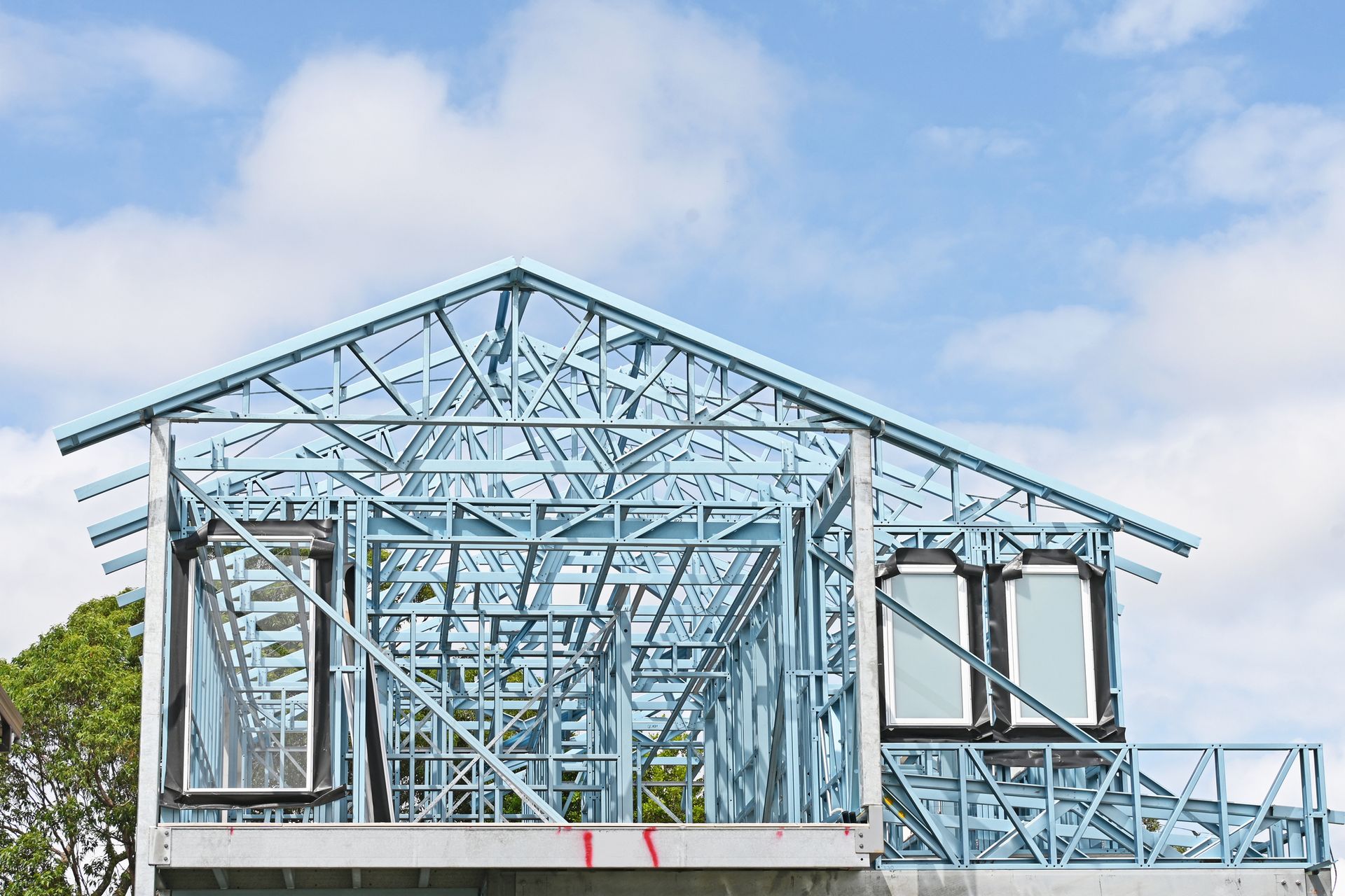 Prefab roof trusses in steel frame home construction under a bright blue sky with white clouds.