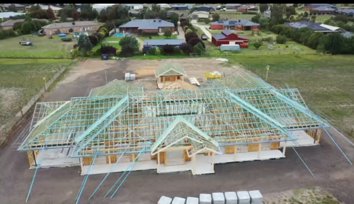 Aerial view of a building under construction, showing wooden roof framing and concrete base in a rural setting.