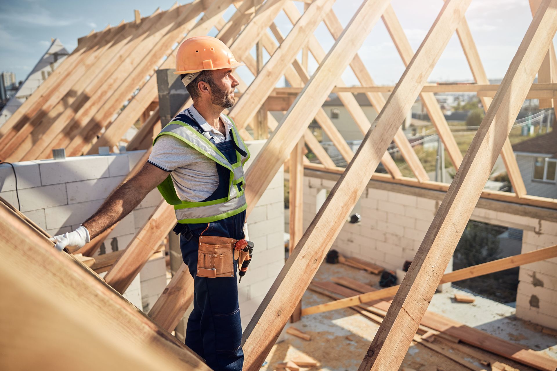 Construction worker on rooftop, wearing a safety vest and hard hat, looking towards the sun.
