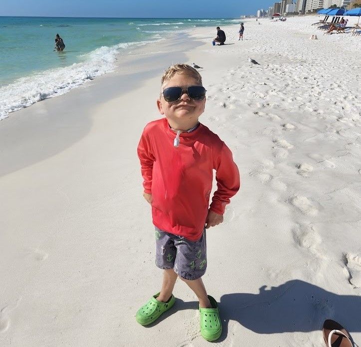 A young boy wearing sunglasses is standing on a beach Cole Kubista