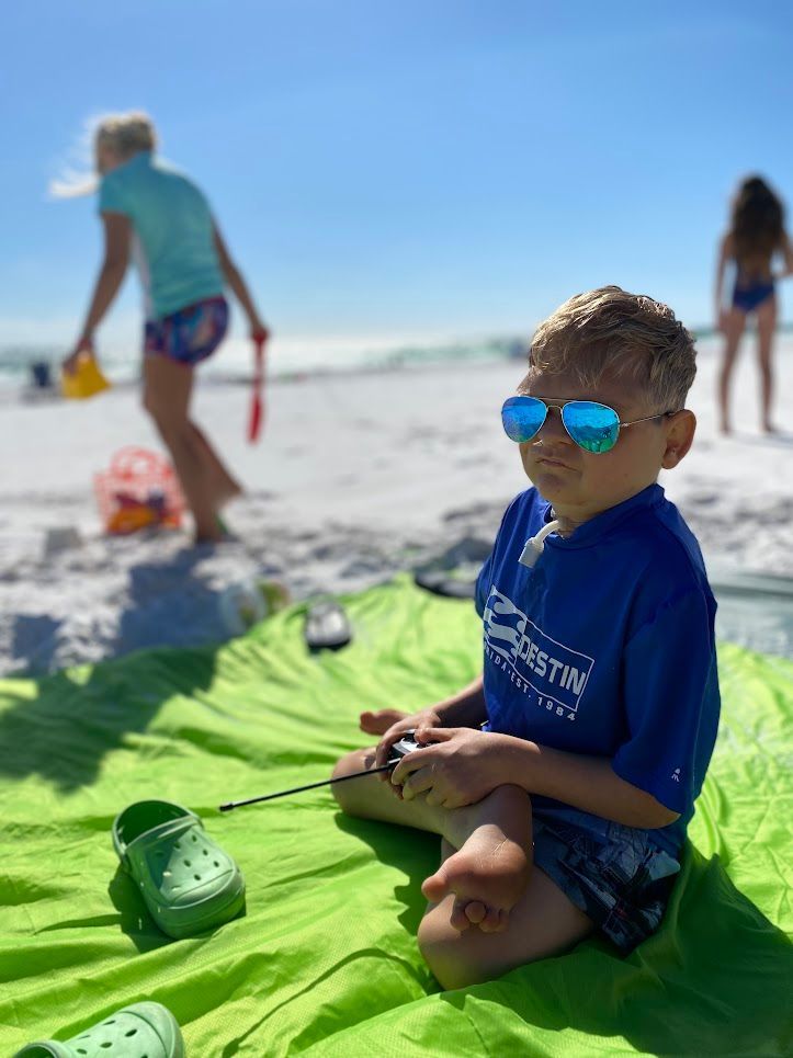 Cole Kubista A young boy wearing sunglasses is sitting on a green towel on the beach.