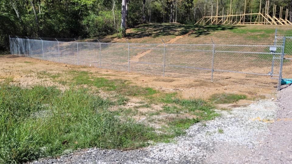 A chain link fence surrounds a dirt field with trees in the background.