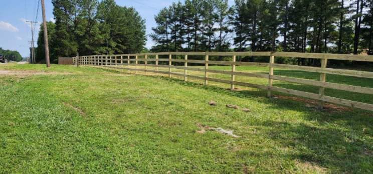 A wooden fence surrounds a grassy field with trees in the background.