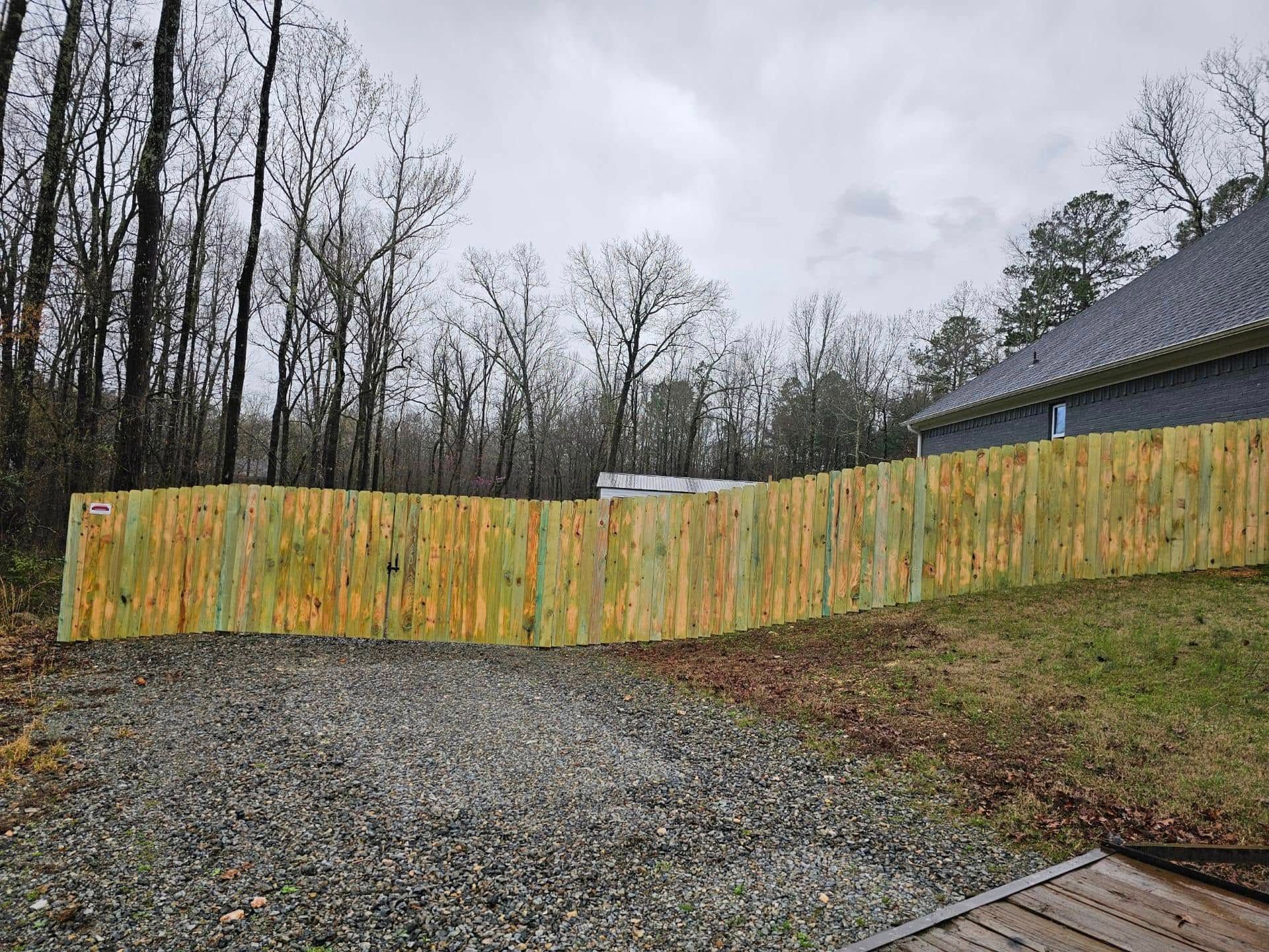 A wooden fence is surrounding a gravel driveway in front of a house.