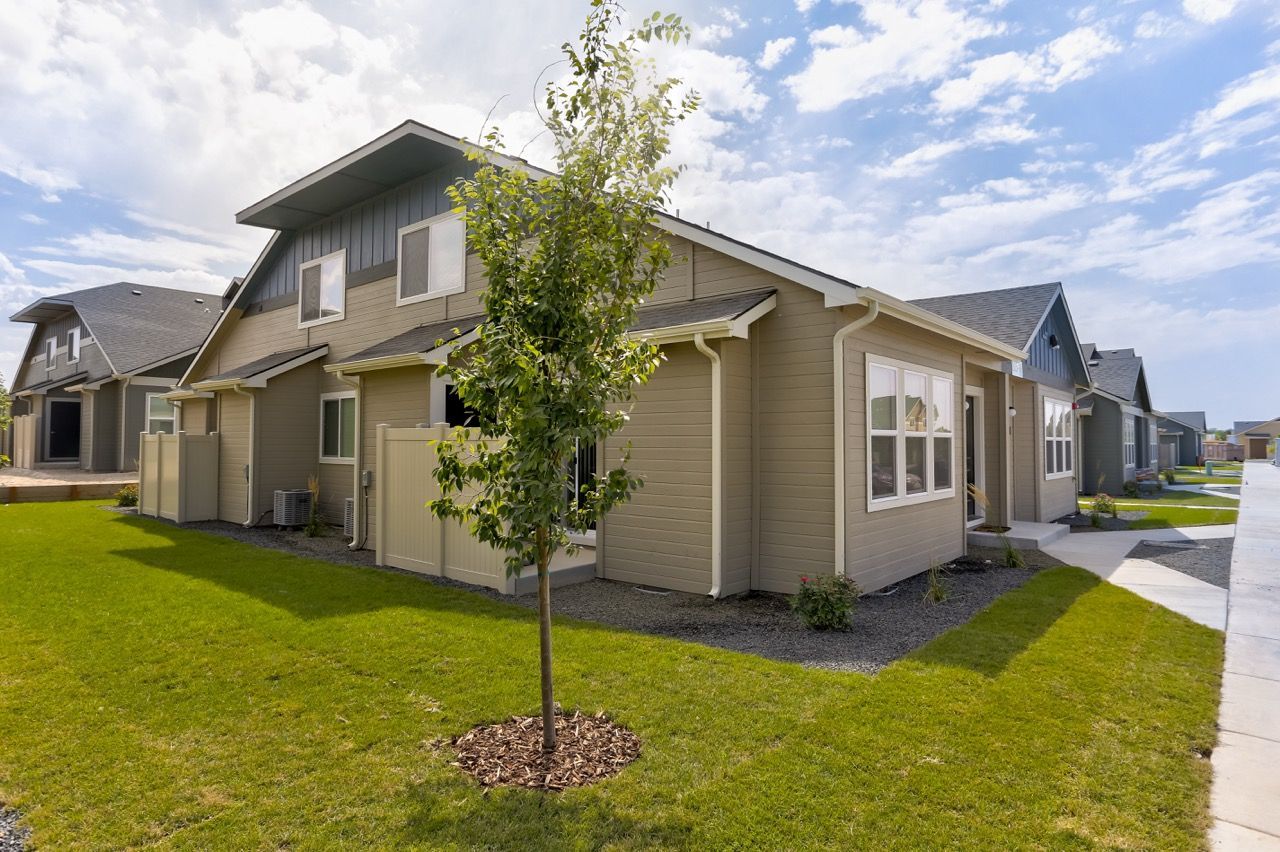 Exterior view of a row of beige townhomes with a young tree in a green lawn and sidewalk.