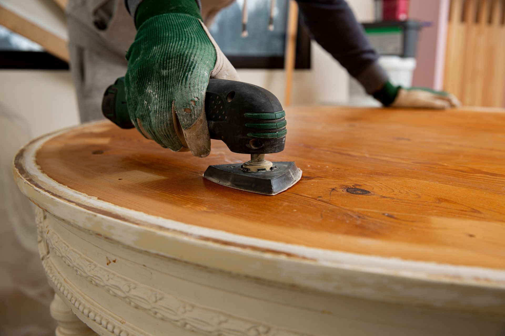 Person sanding a wooden table with an electric sander, wearing gloves.
