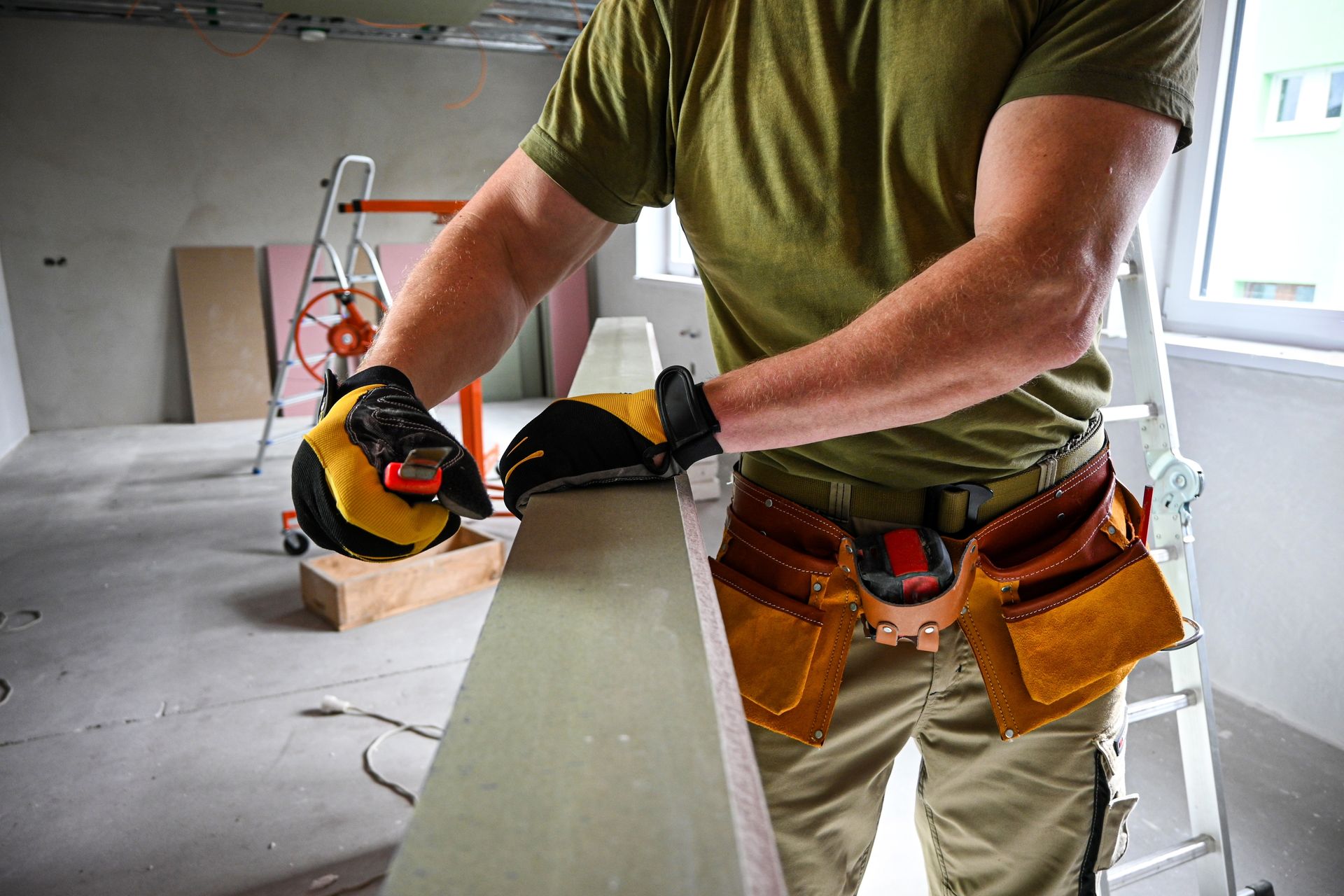 Person wearing gloves sanding a wooden plank indoors, with tool belt.