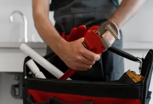 Plumber holding a red pipe wrench, reaching into toolbox containing pipes and gloves, near a sink.