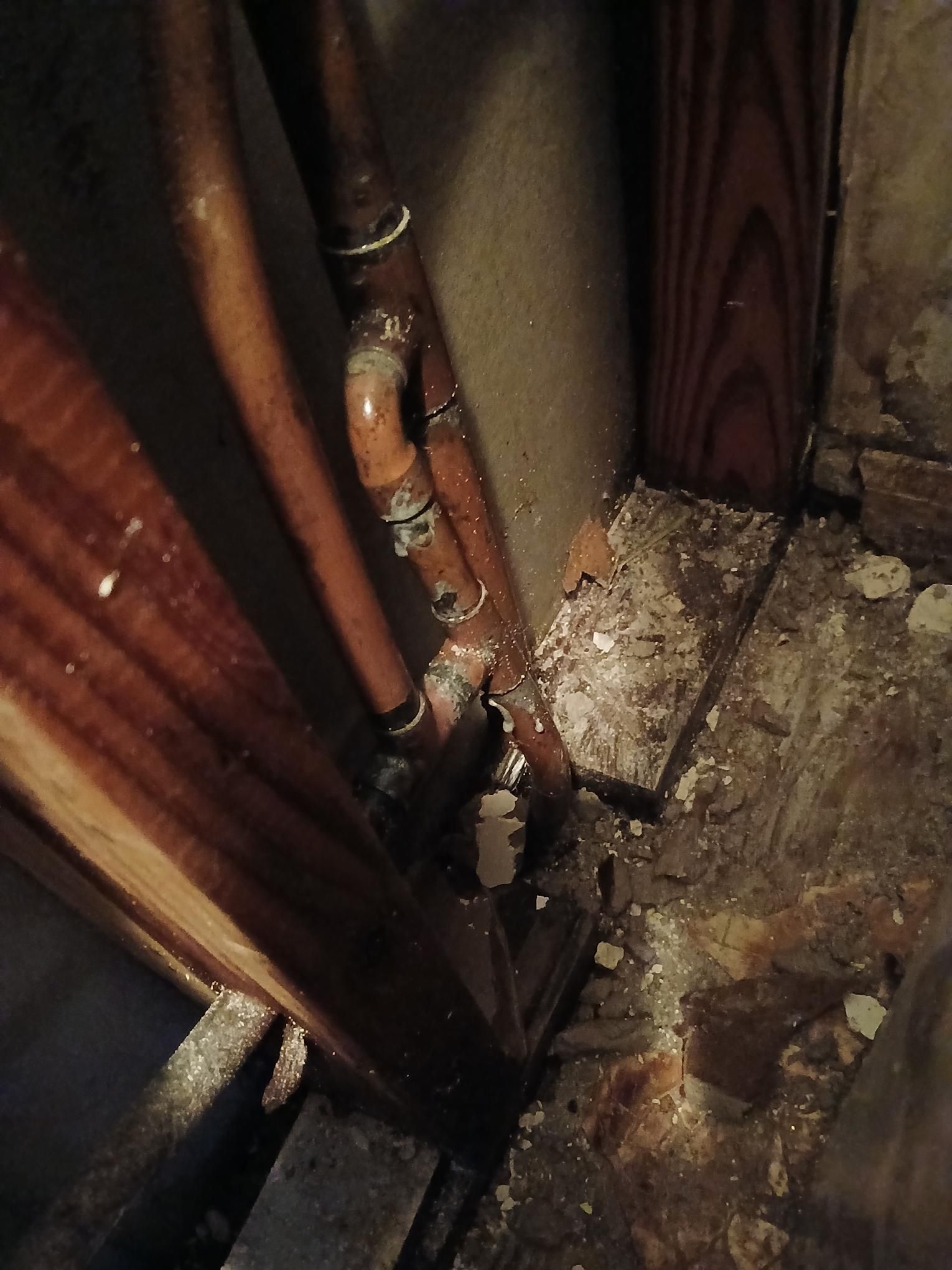 Copper pipes with water damage in a corner of a basement, next to wooden beams and concrete.