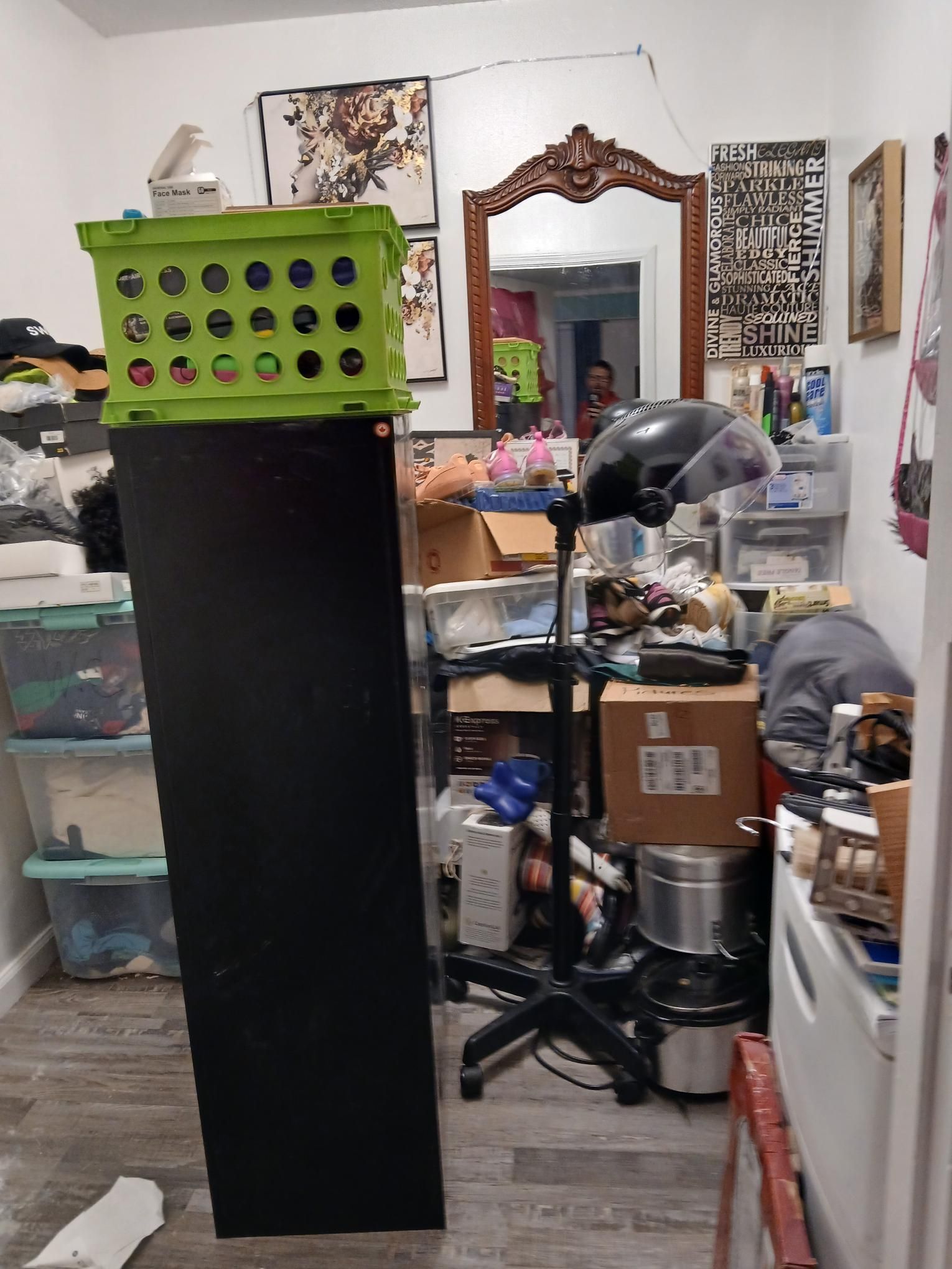 Cluttered room with a black cabinet topped by a green crate. A mirror, hair dryer, and various items are present.