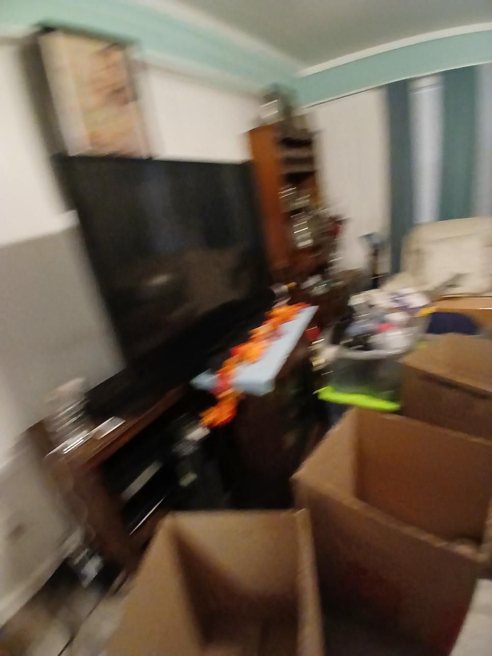 Living room with a large TV, boxes, and furniture, appears cluttered.