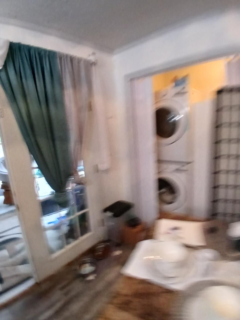Interior shot with a window, washer/dryer, and a table with dishes. Teal and white curtains, wooden floor.