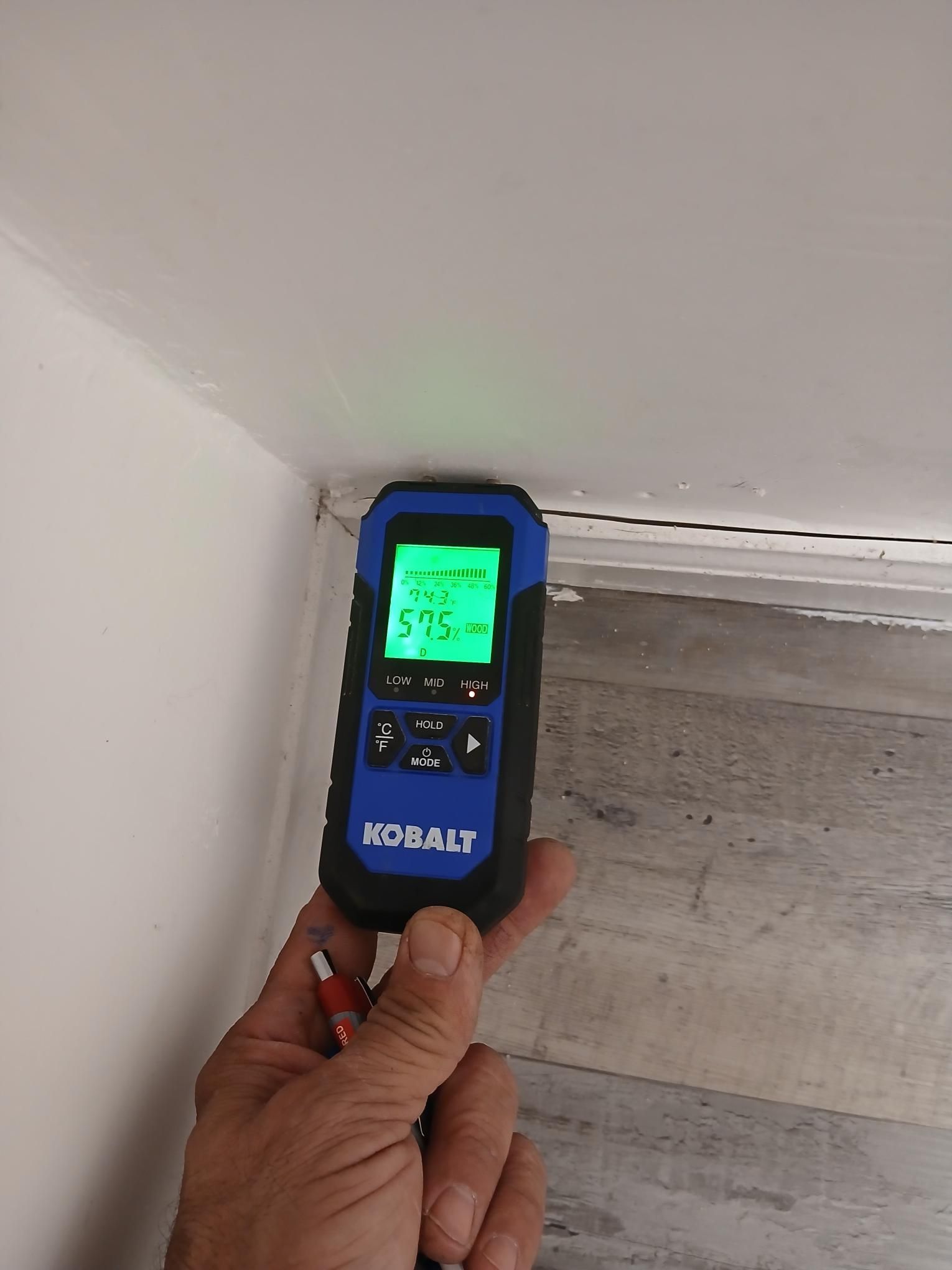 Hand holding a moisture meter against a white wall and ceiling corner, showing a green reading on the screen.