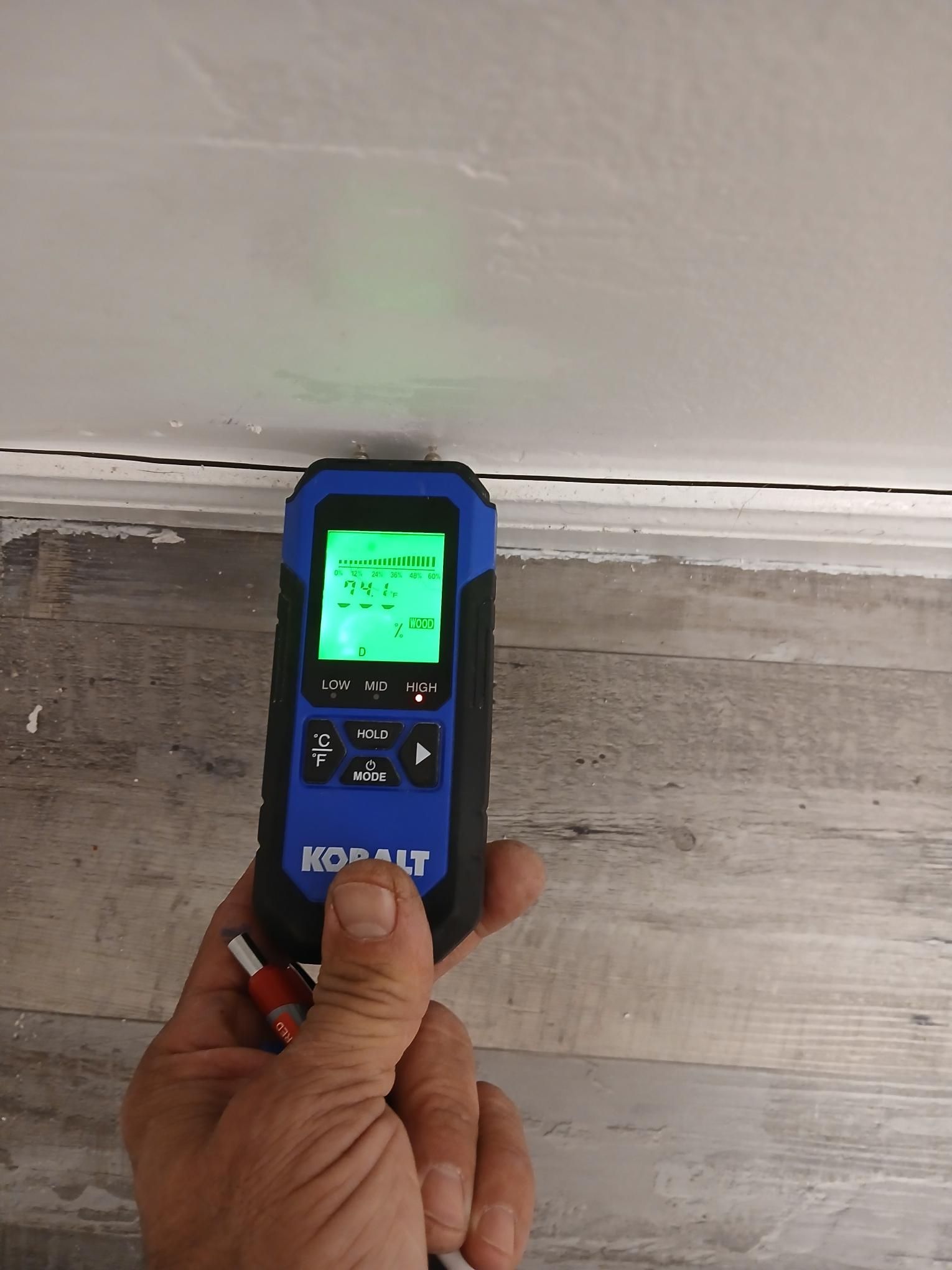 Person holding a blue Kobalt stud finder against a wall. The device displays green numbers.