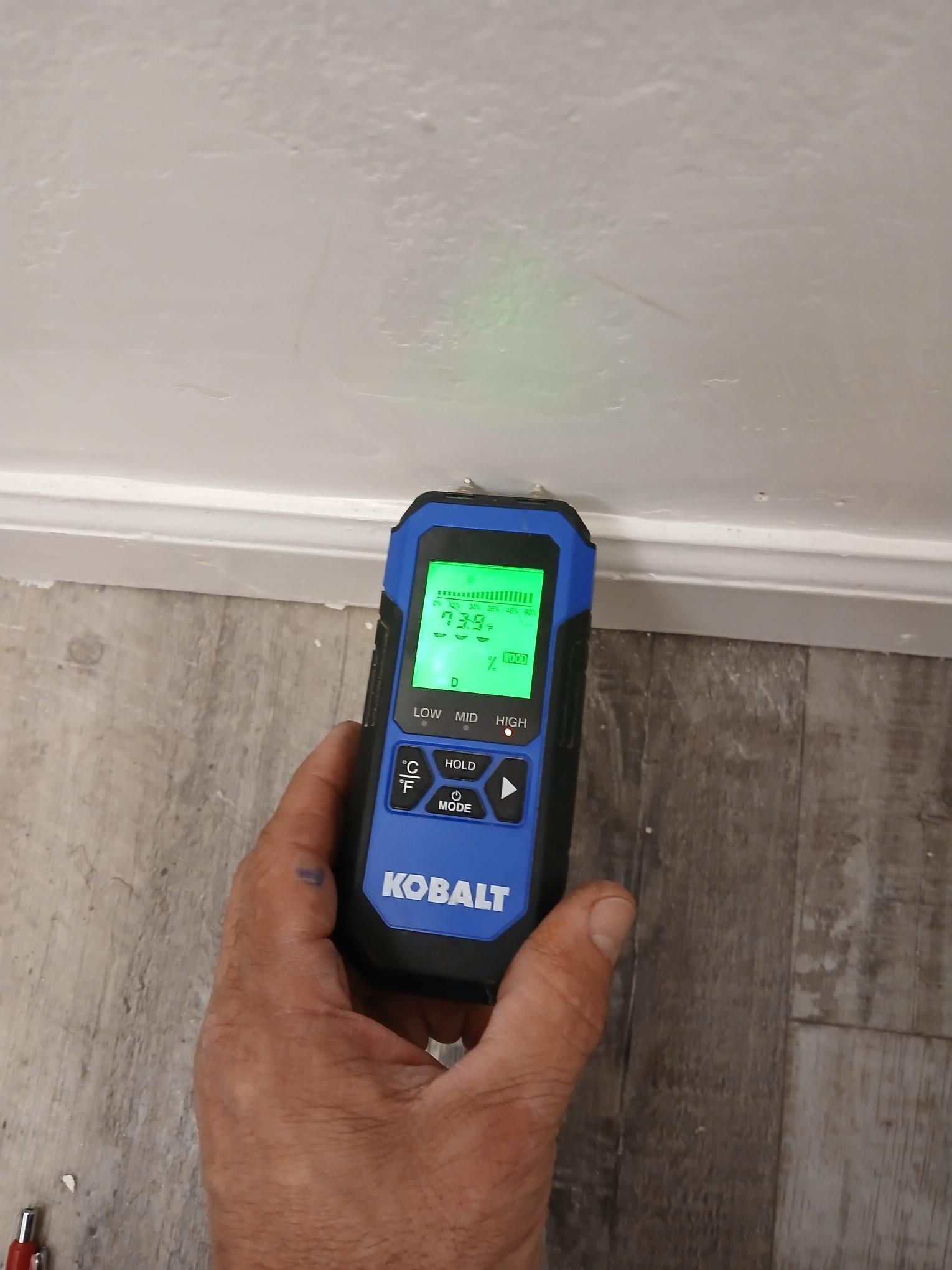 A person holds a blue and black Kobalt moisture meter against a white wall near a floor.