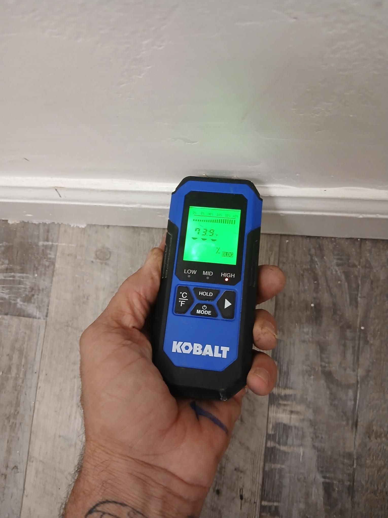 Hand holding a blue and black stud finder near a white wall and baseboard, with a green display.