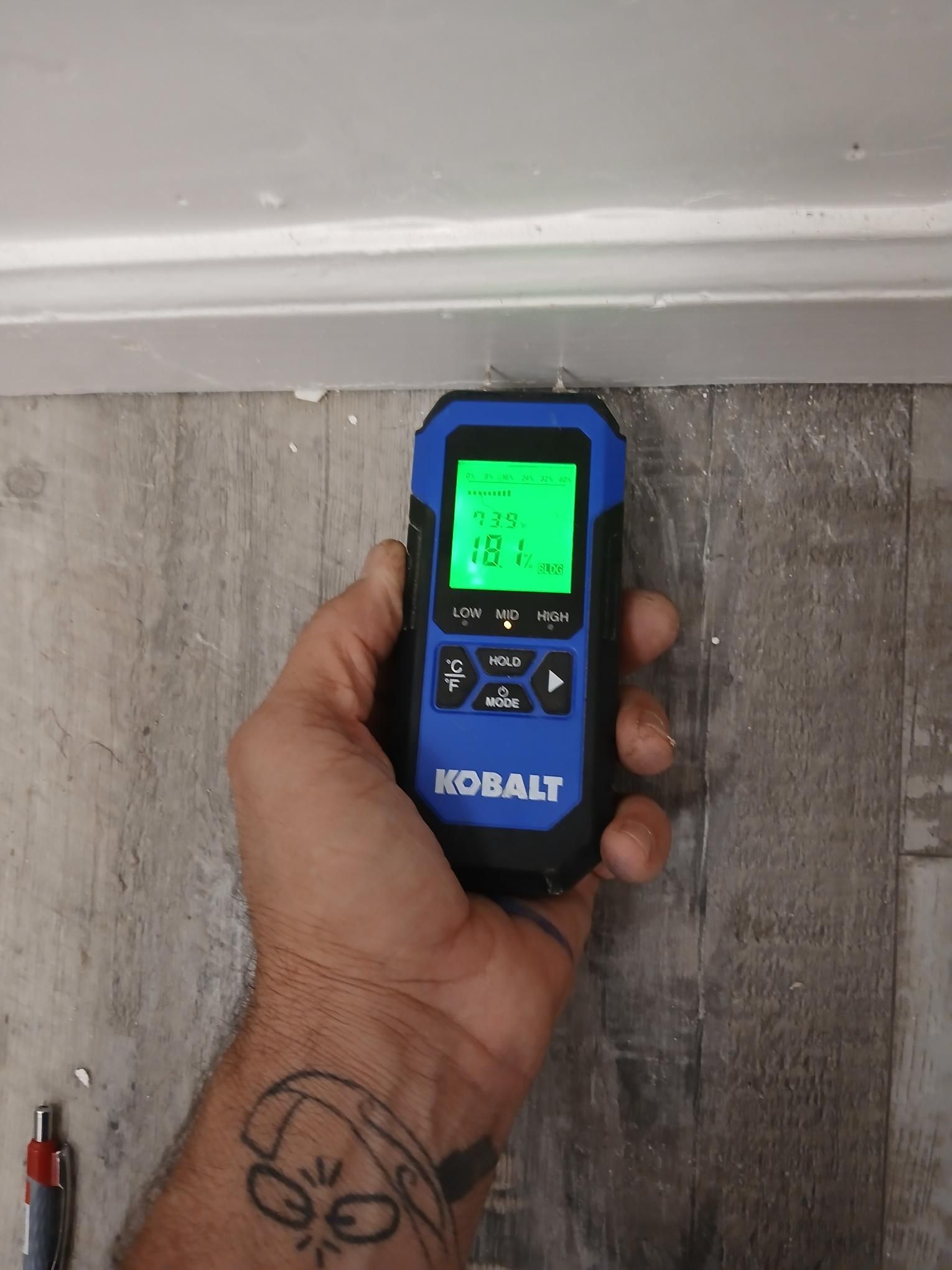 Hand holding a blue and black moisture meter near a white baseboard and gray flooring; meter displays 19.1%.
