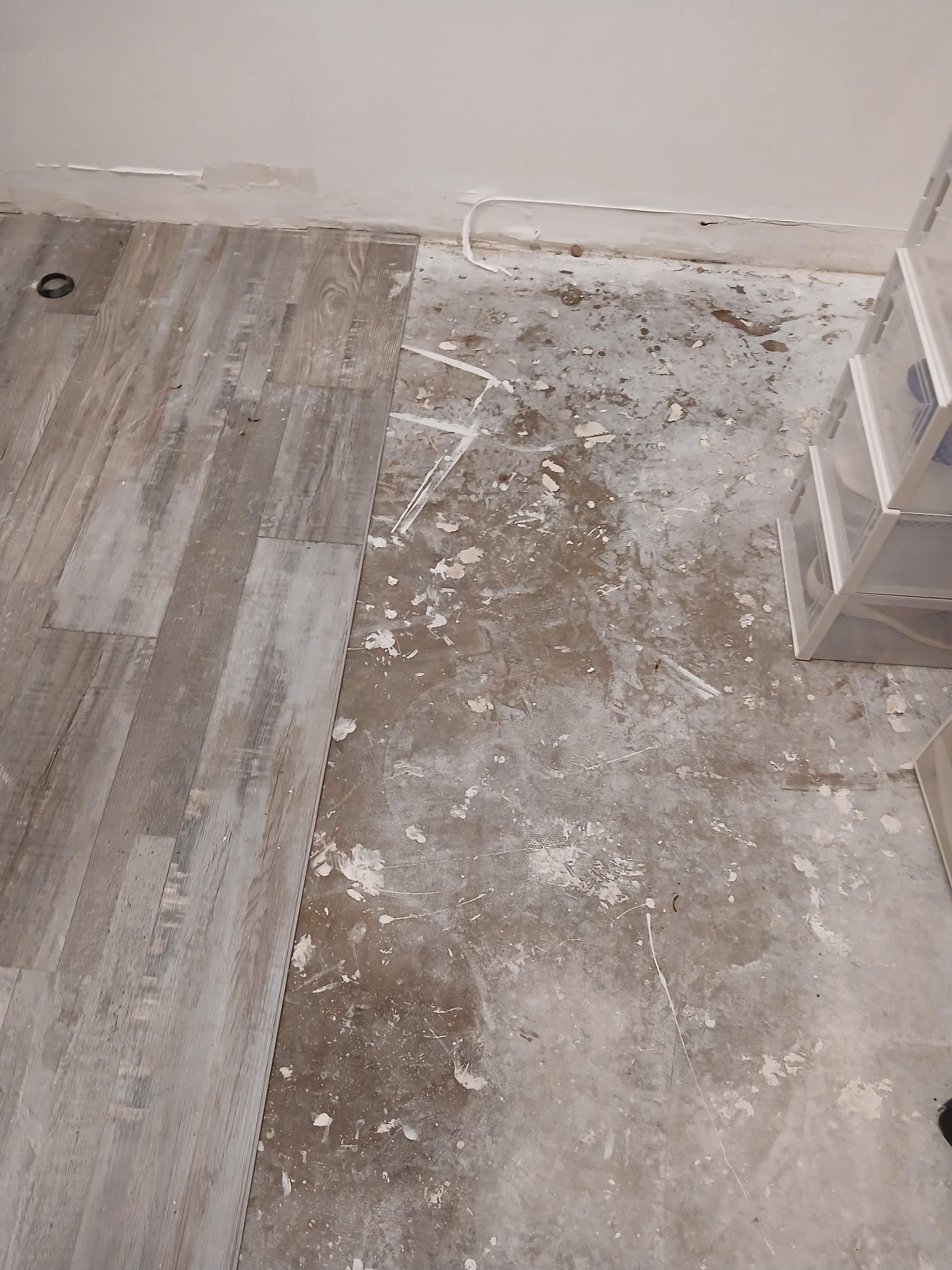 Damaged flooring with white debris, next to a white wall and storage unit.