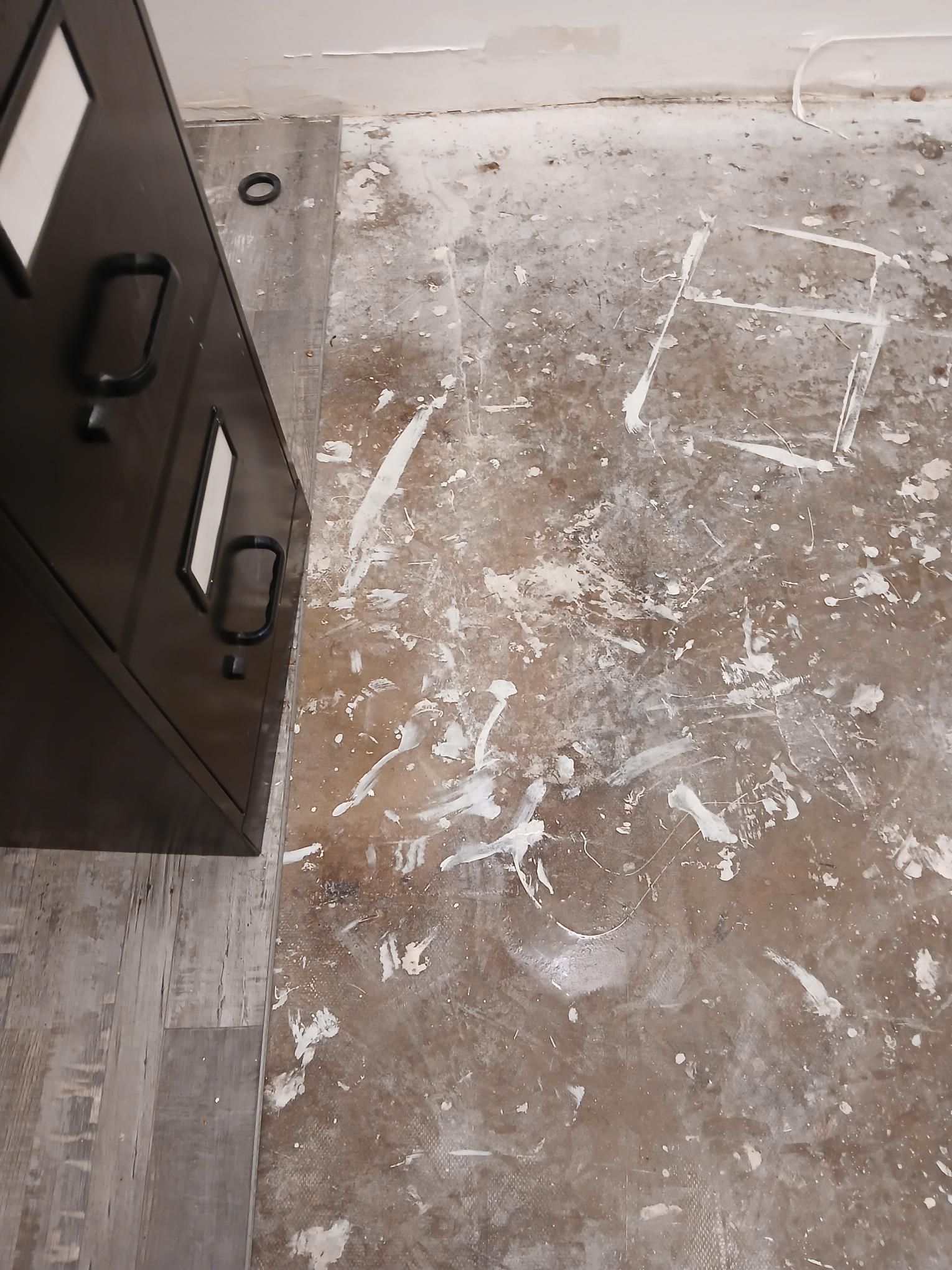 Close-up of a floor with white paint splatters and a dark cabinet.