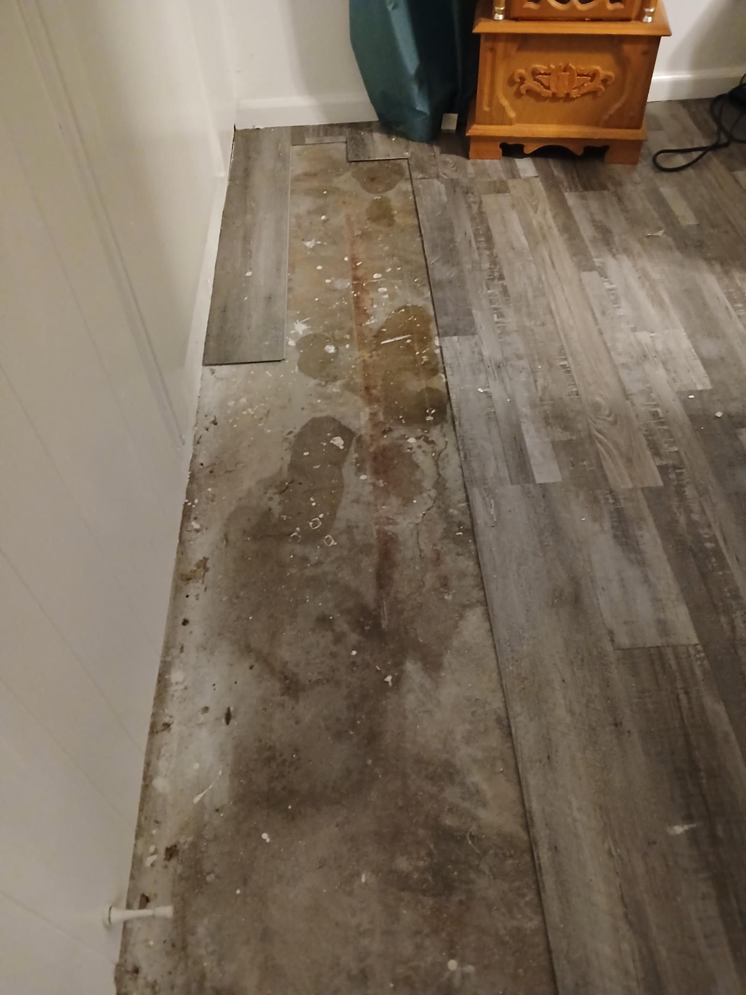 Damaged gray vinyl flooring with water stains, next to a white wall and wood furniture.