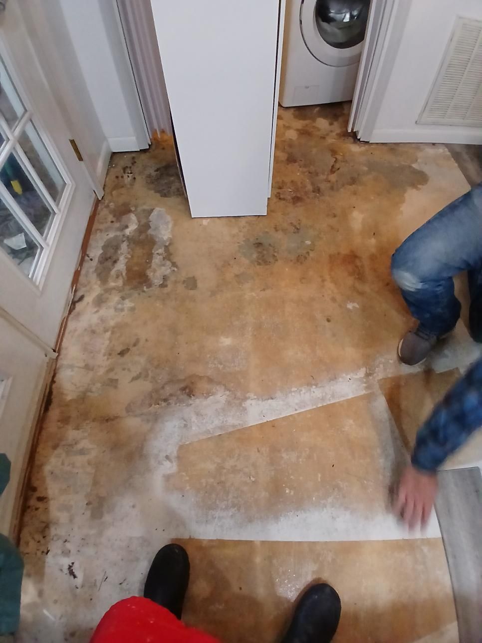 Water-damaged floor with two people; one kneels, the other reaches. A washing machine and white cabinet are visible.