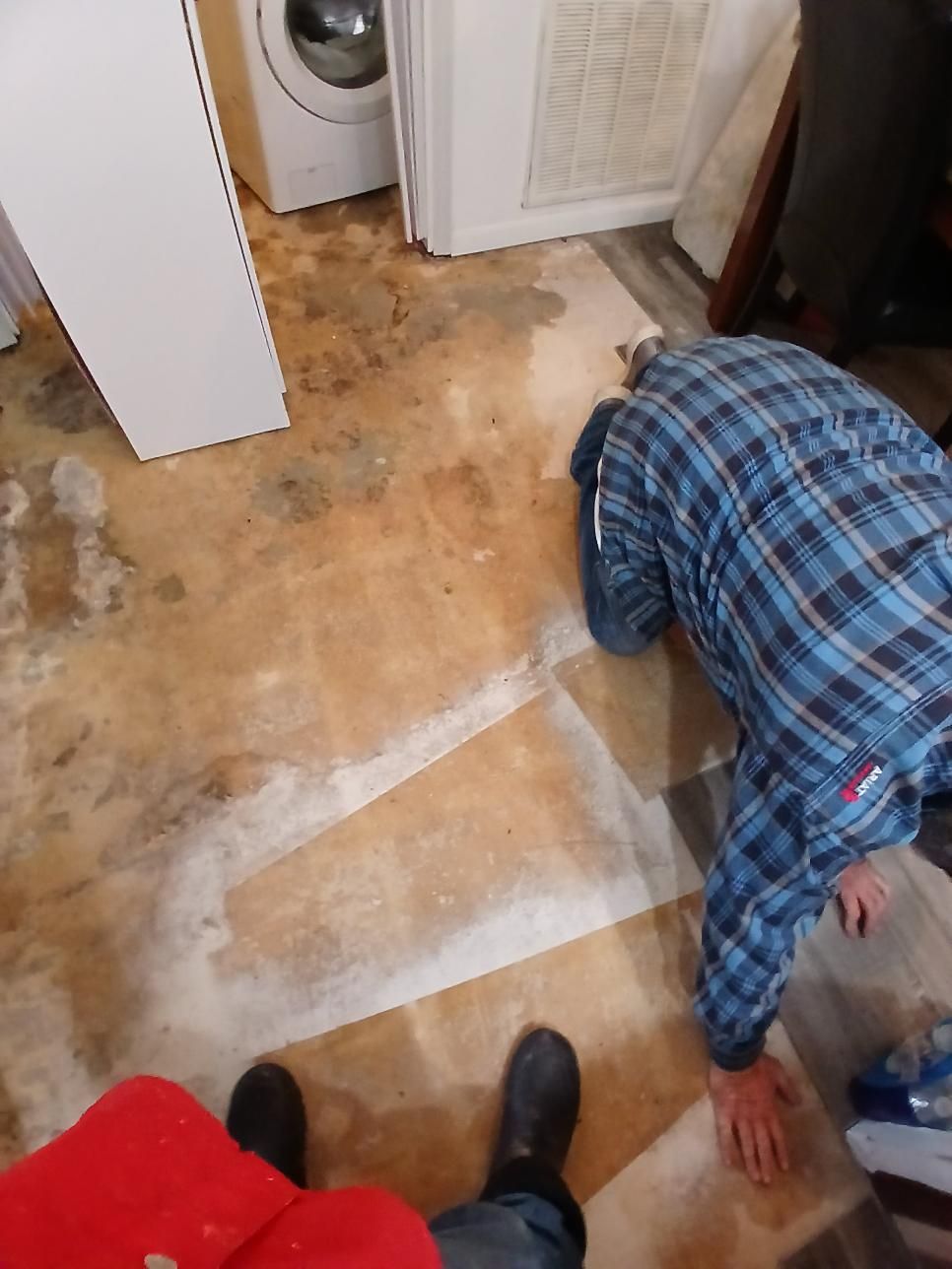 Person on hands and knees near a damaged floor. White appliance and cabinet visible. Water damage evident.