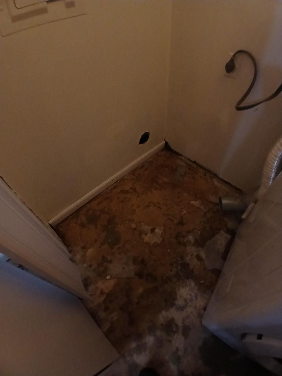Laundry room with damaged flooring and a dryer. Beige walls, visible dryer vent, and electrical cord.