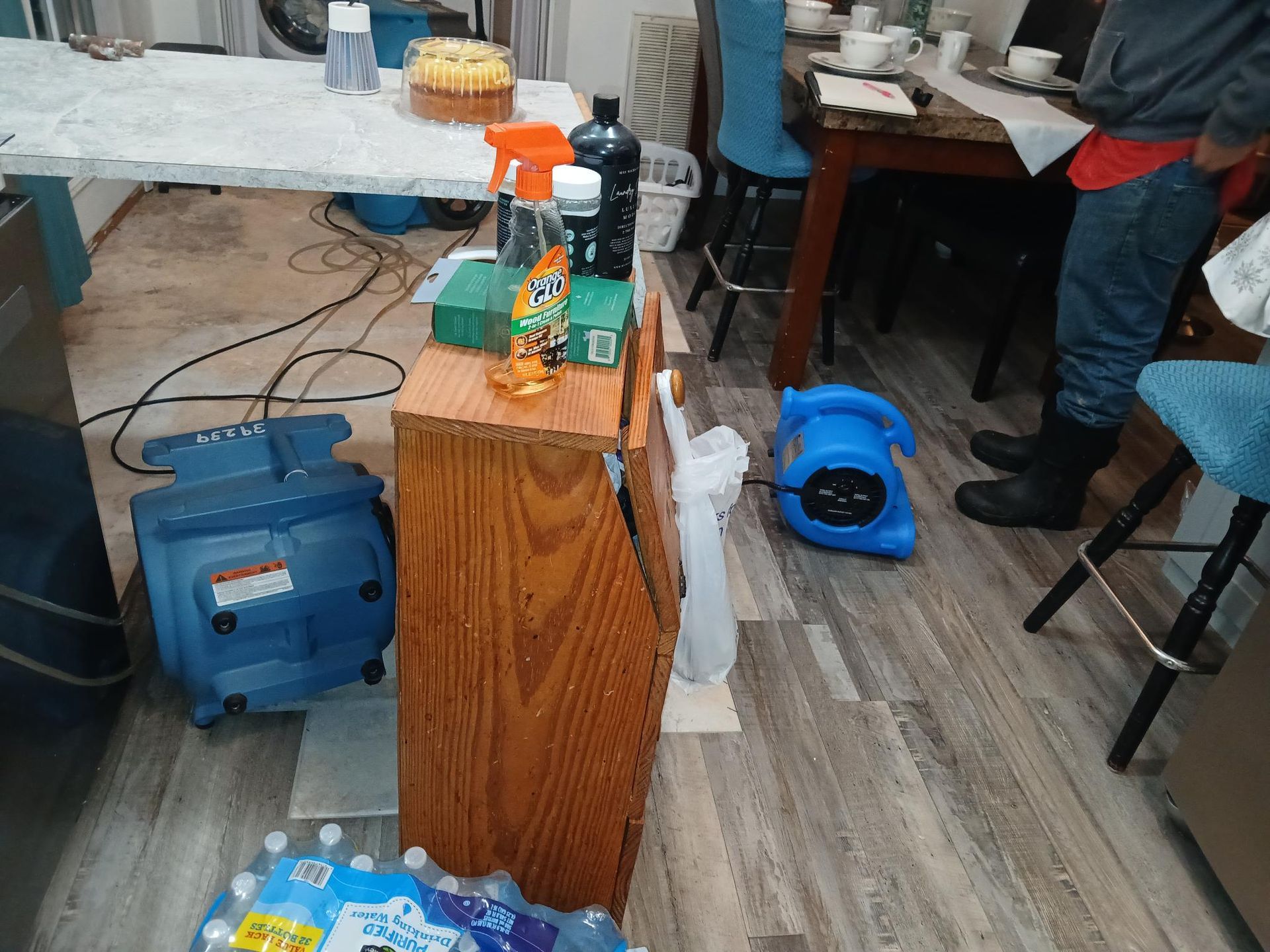 A room with a water damage restoration in progress. A person stands near a table and chair. Several drying fans are in use.