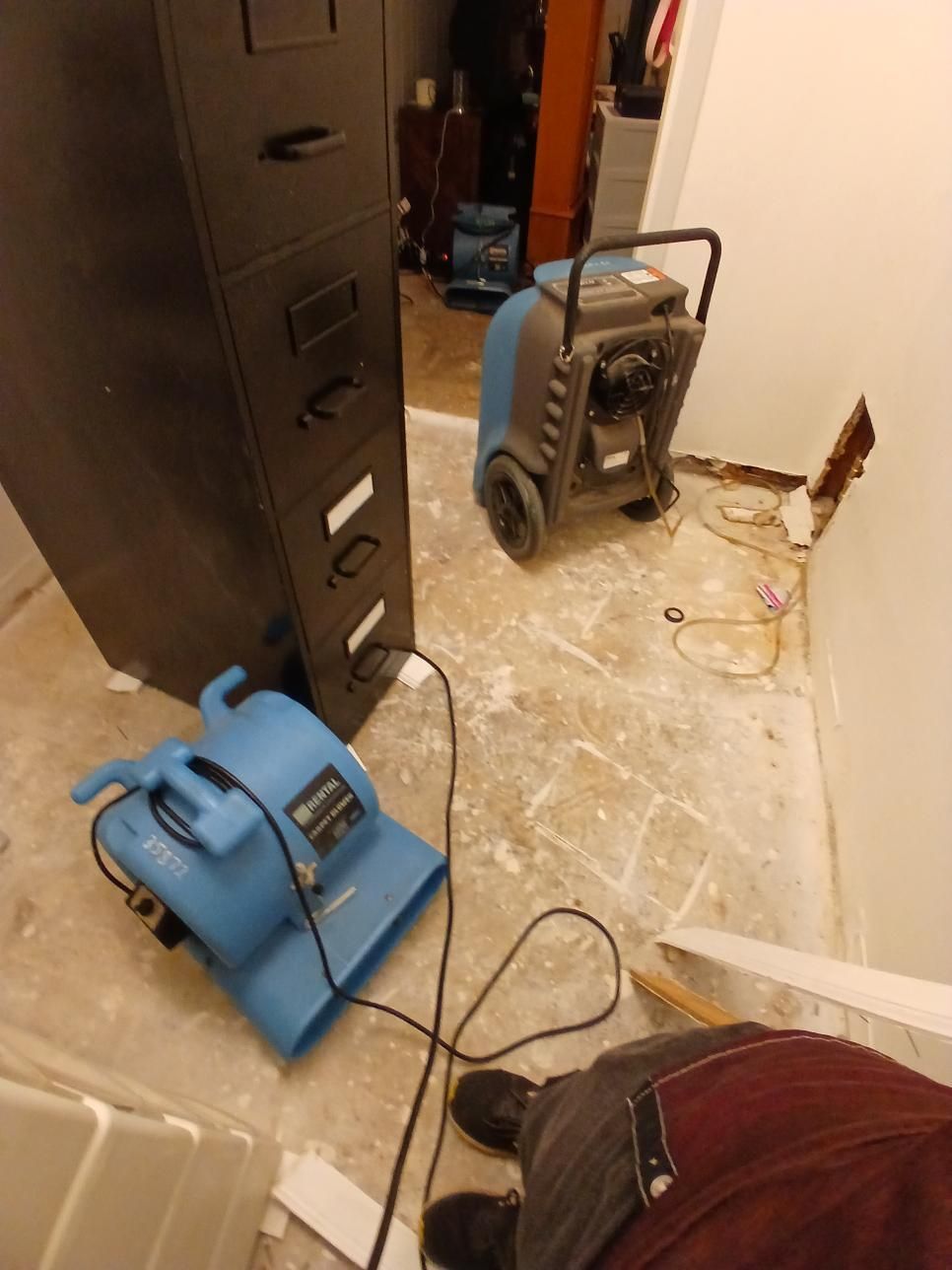 A blower and dehumidifier in a room with a damaged wall, by a file cabinet.