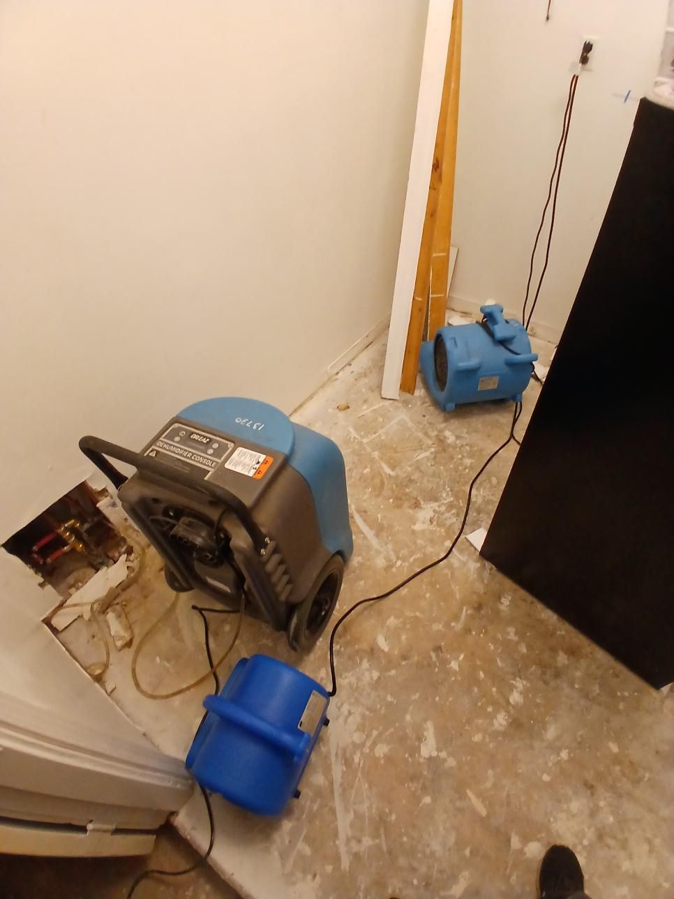 Dehumidifiers and a fan in a room with water damage. Beige walls, gray flooring. Electrical components exposed.