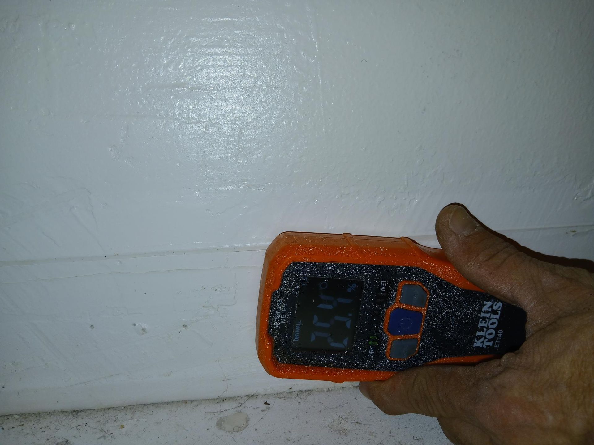 Hand holding a moisture meter against a white painted wall, reading 15.4%.