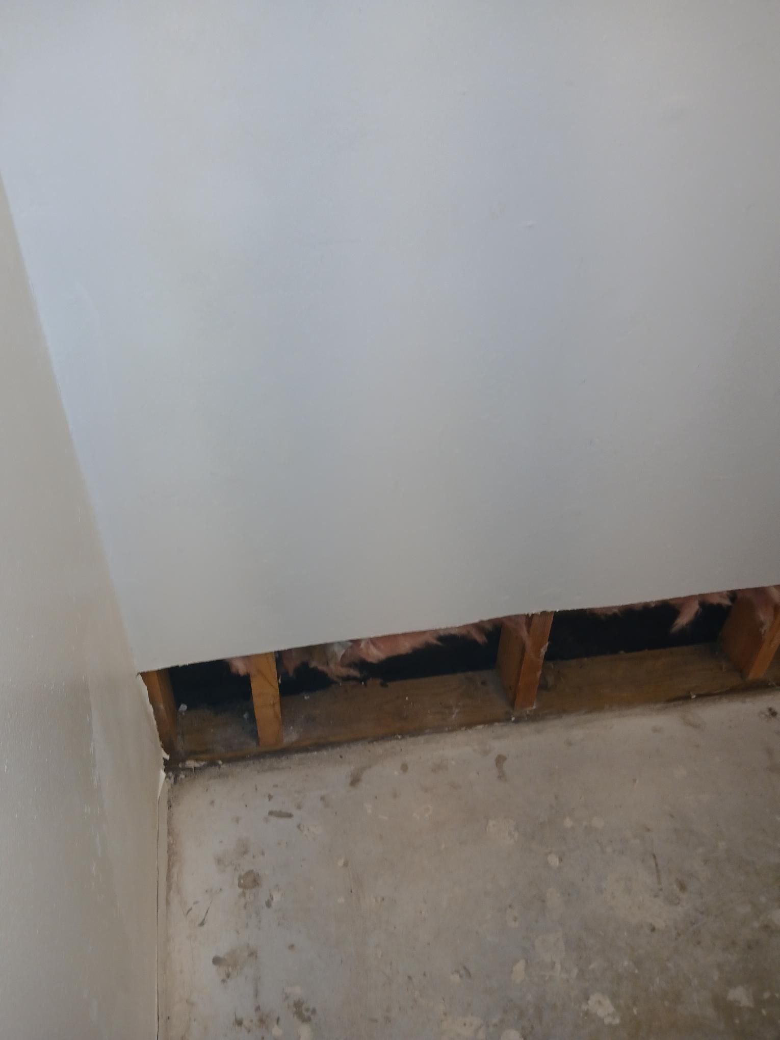 Hole in the wall showing exposed wooden beams and insulation, concrete floor, and white walls.