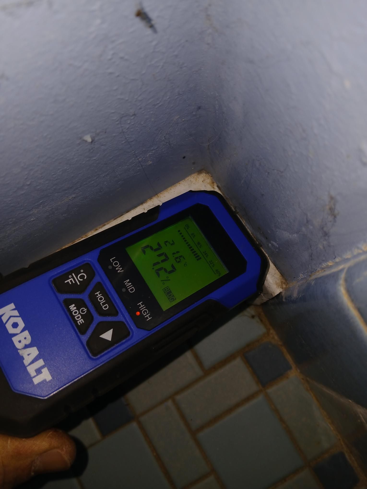 Blue moisture meter reading 27.2 in corner of bathroom, tiles visible.