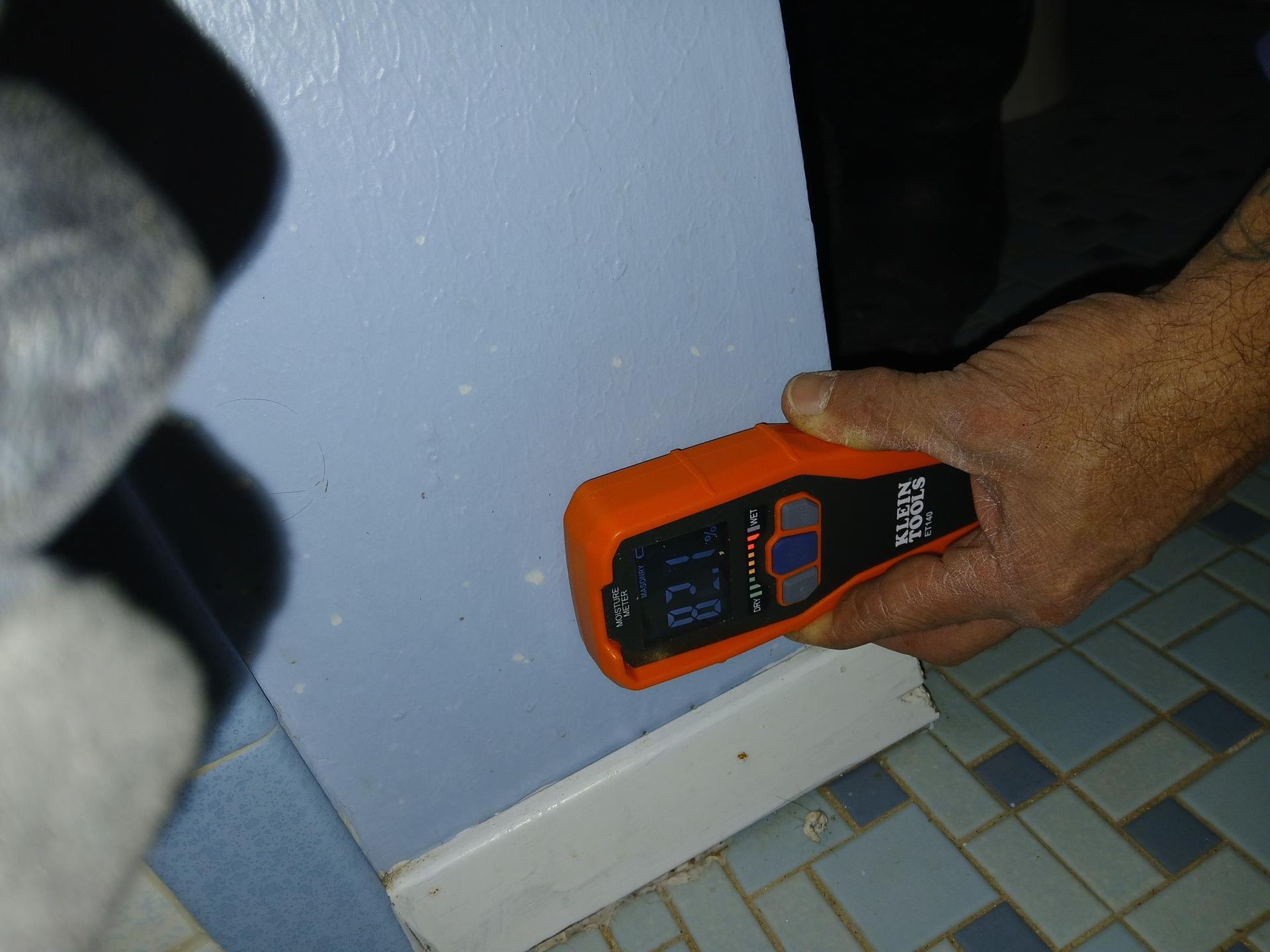 A person using an orange stud finder on a light blue wall near a white baseboard and blue tile floor.