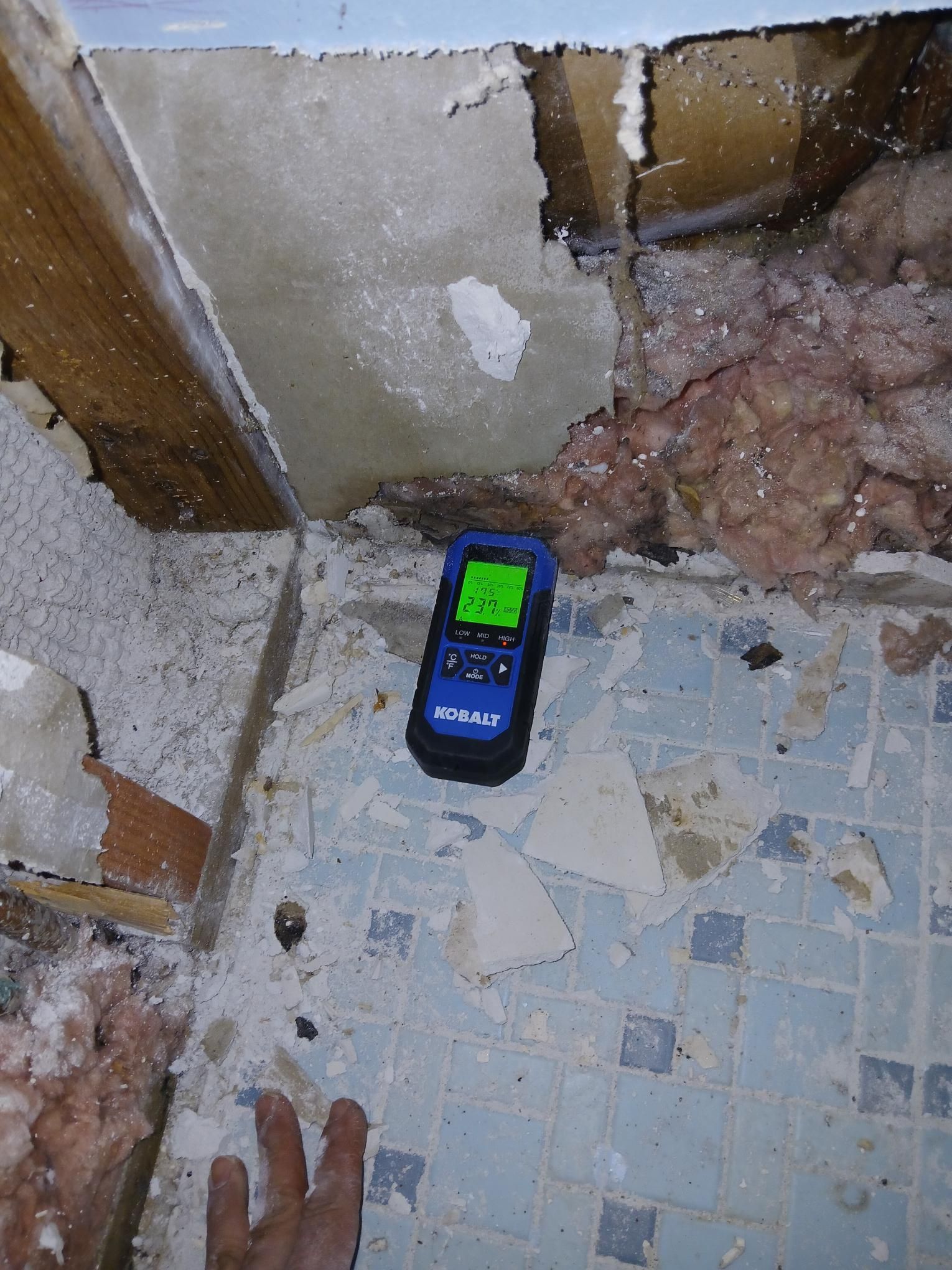 A moisture meter on blue tile floor near damaged walls. Hand is visible.
