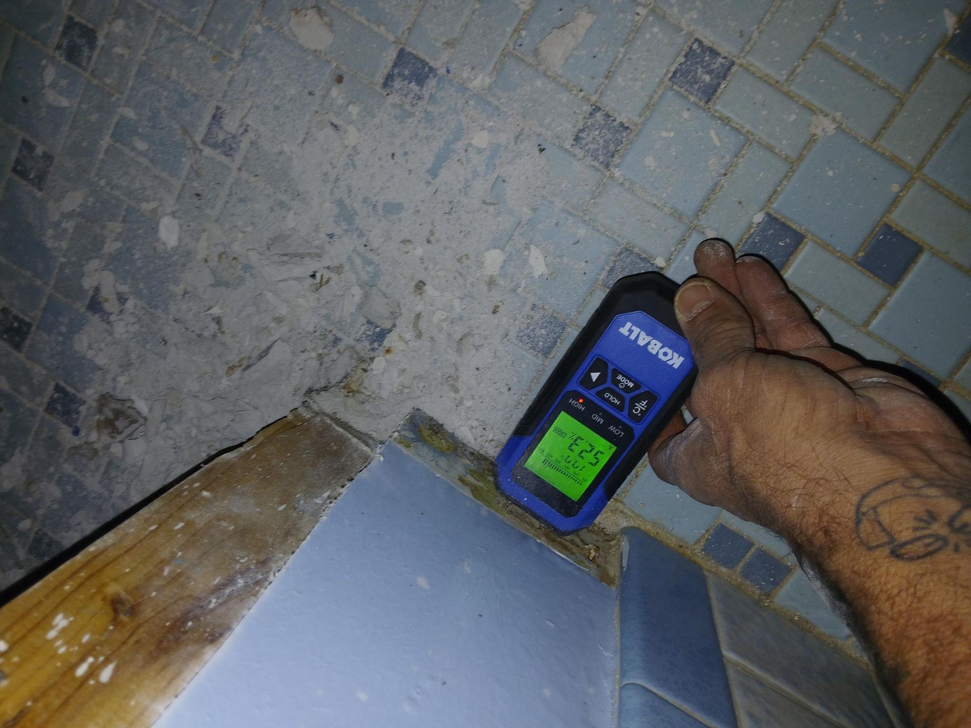 Hand holding a moisture meter on damaged bathroom floor tiles, reading 27.5.