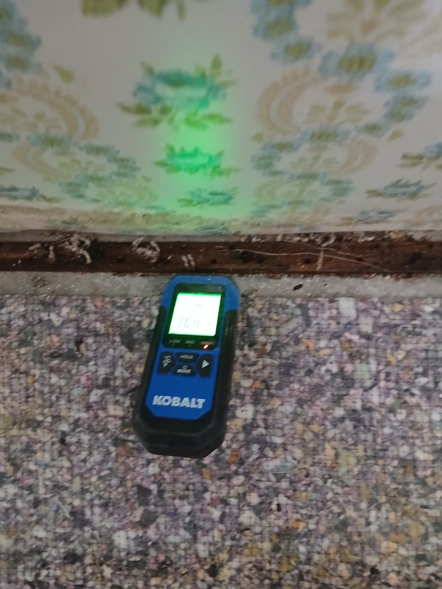 A stud finder is used on a wall with patterned wallpaper and carpet.