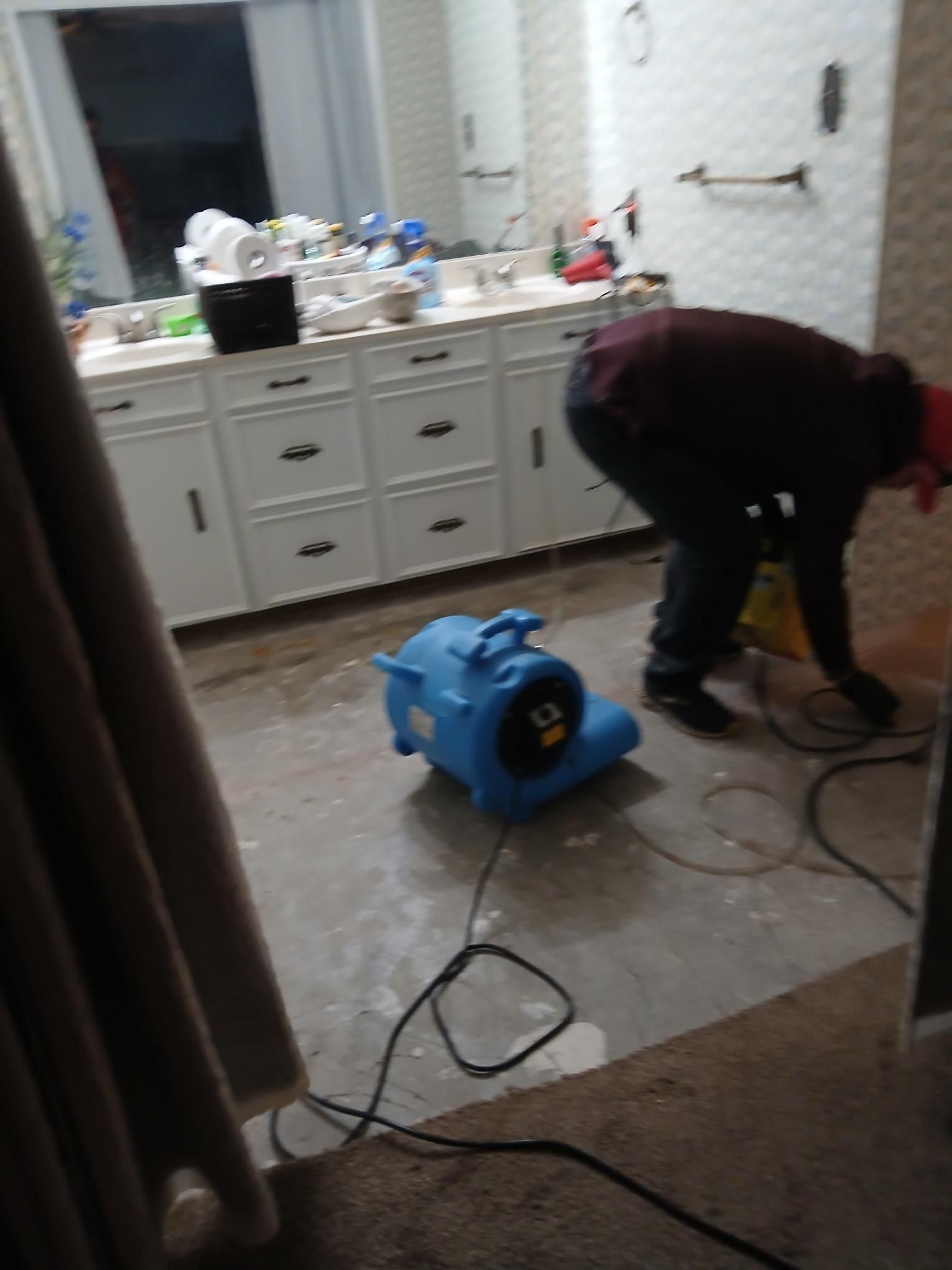 A person using a blue air mover to dry a flooded bathroom floor. White cabinets in the background.