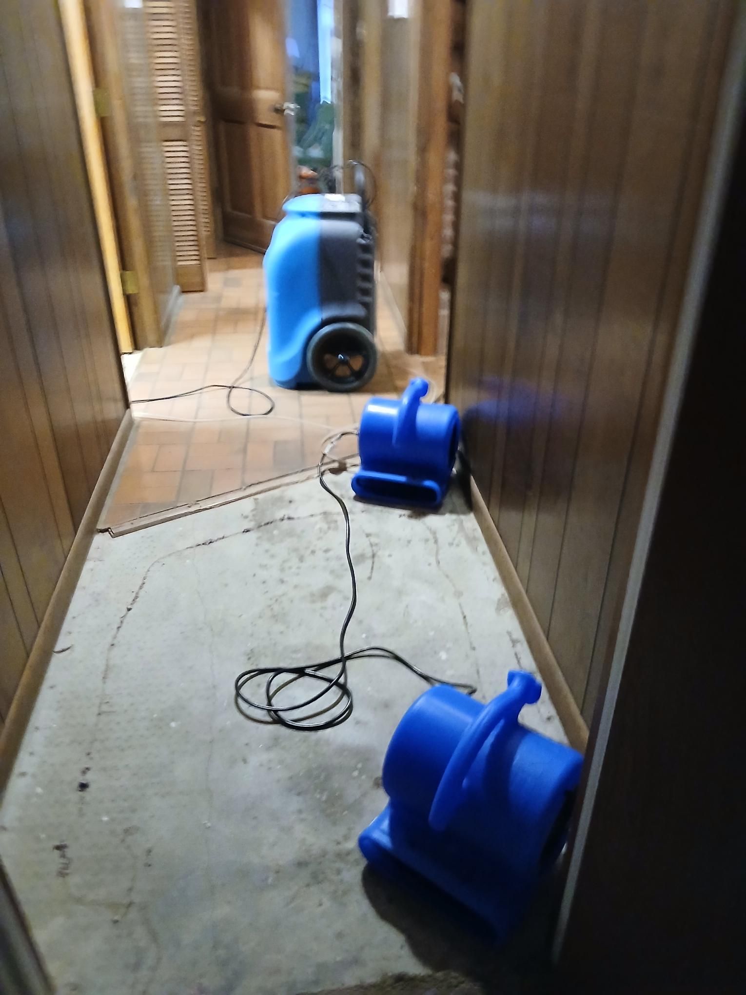 Blue dehumidifier and fans drying a hallway with exposed concrete floor. Wooden walls and doors.