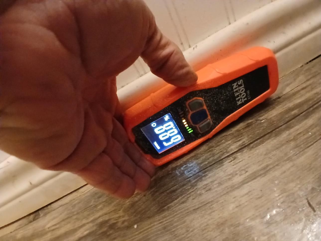 Hand holding orange moisture meter on wood floor near white molding. Display shows a reading of 88.9.