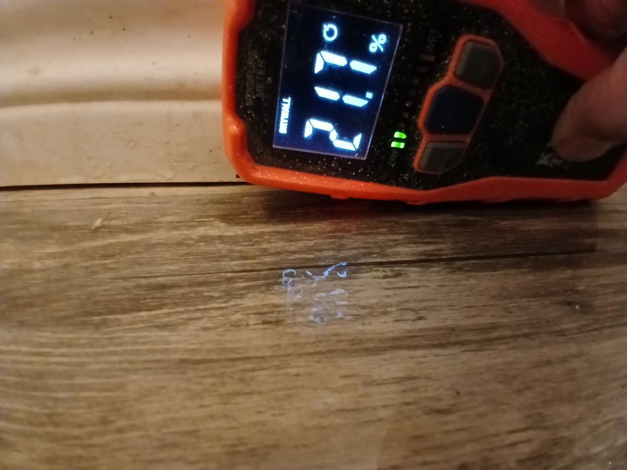 A hand holding a temperature gauge over a light-colored wood floor with a small wet spot. The gauge reads 21°C.