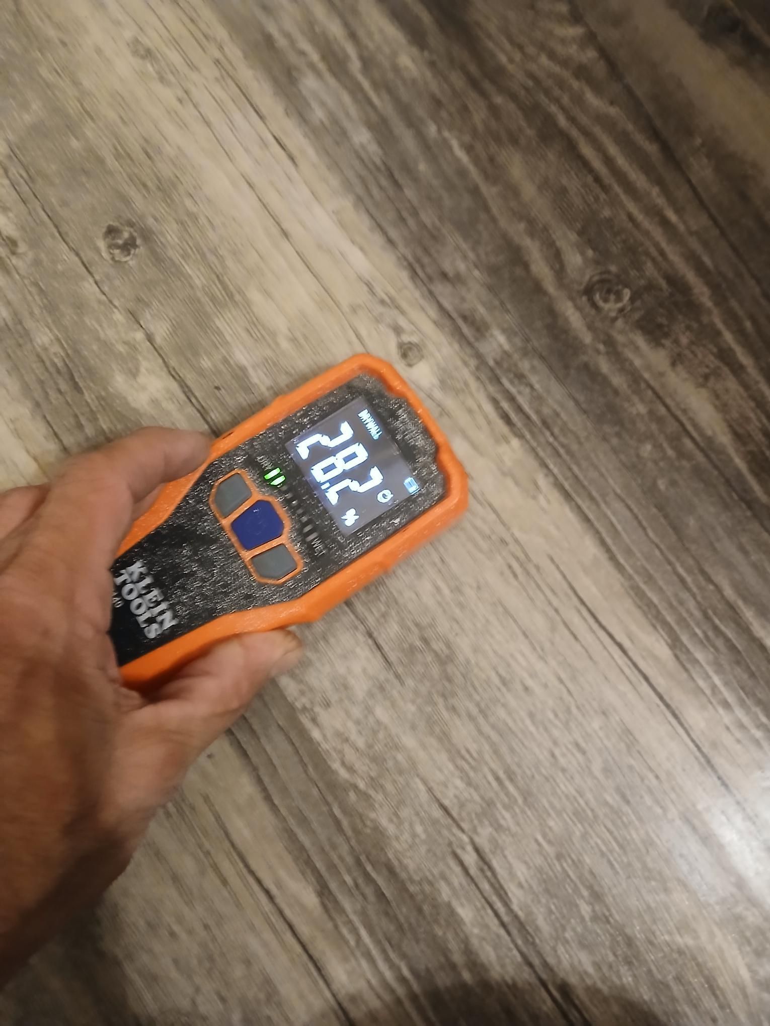 Hand holding an orange digital temperature meter on a wood-look floor, displaying 28.2 degrees.