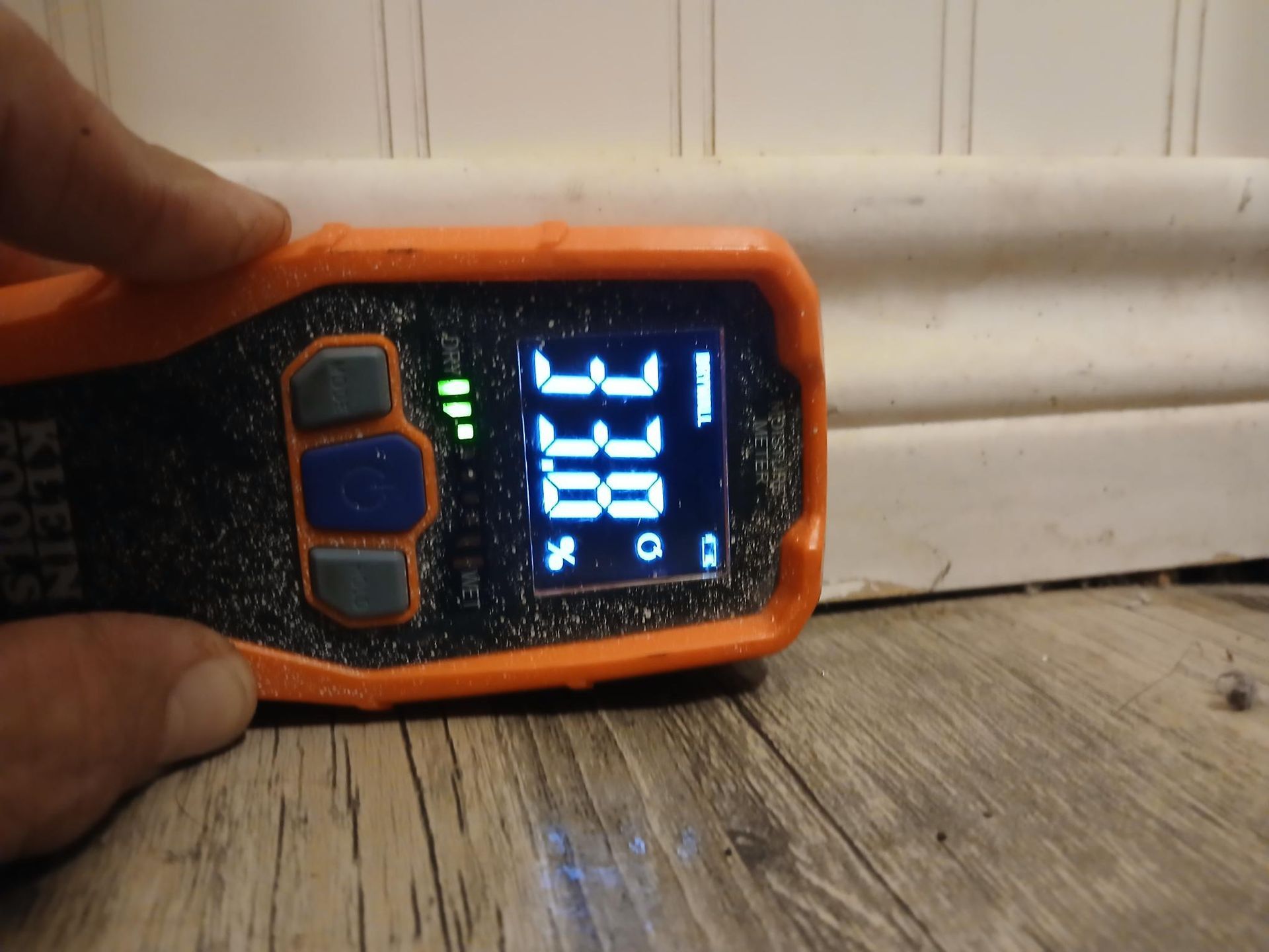 A hand holds a Klein Tools moisture meter, reading 33.8 near a baseboard and wood floor.